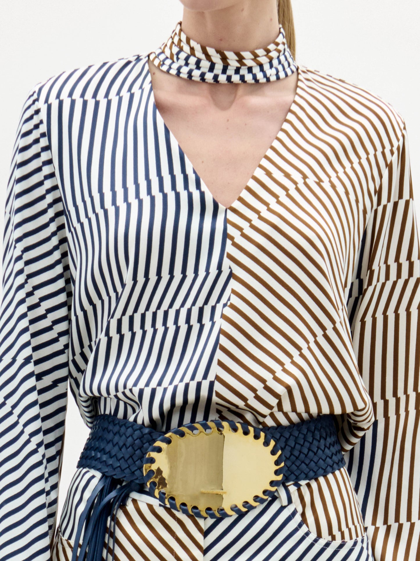 Woman wearing the Jasper Blouse, a long-sleeved, blue and beige striped top, paired with a matching choker and a wide belt with a large gold buckle, stands against a white background.