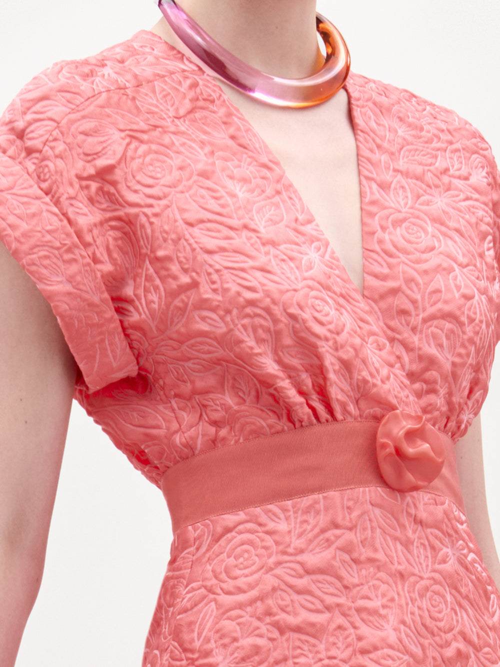 A woman models the Metaponto Dress, a coral textured midi with a fitted waist, styled with a bright pink clutch, brown sandals, and a simple necklace against a plain white background.