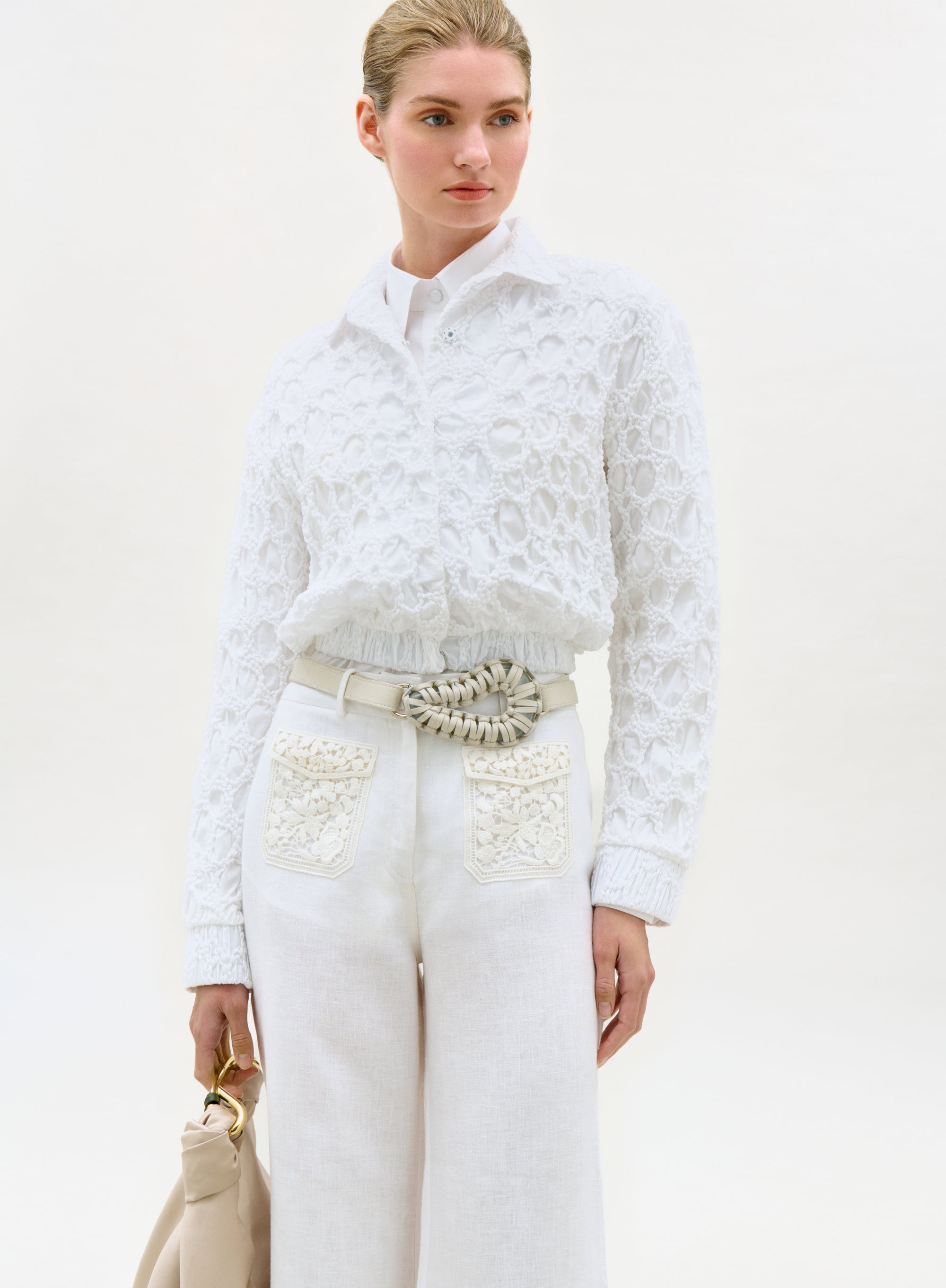 Woman wearing a white lace top and pants with a beige handbag on a light background