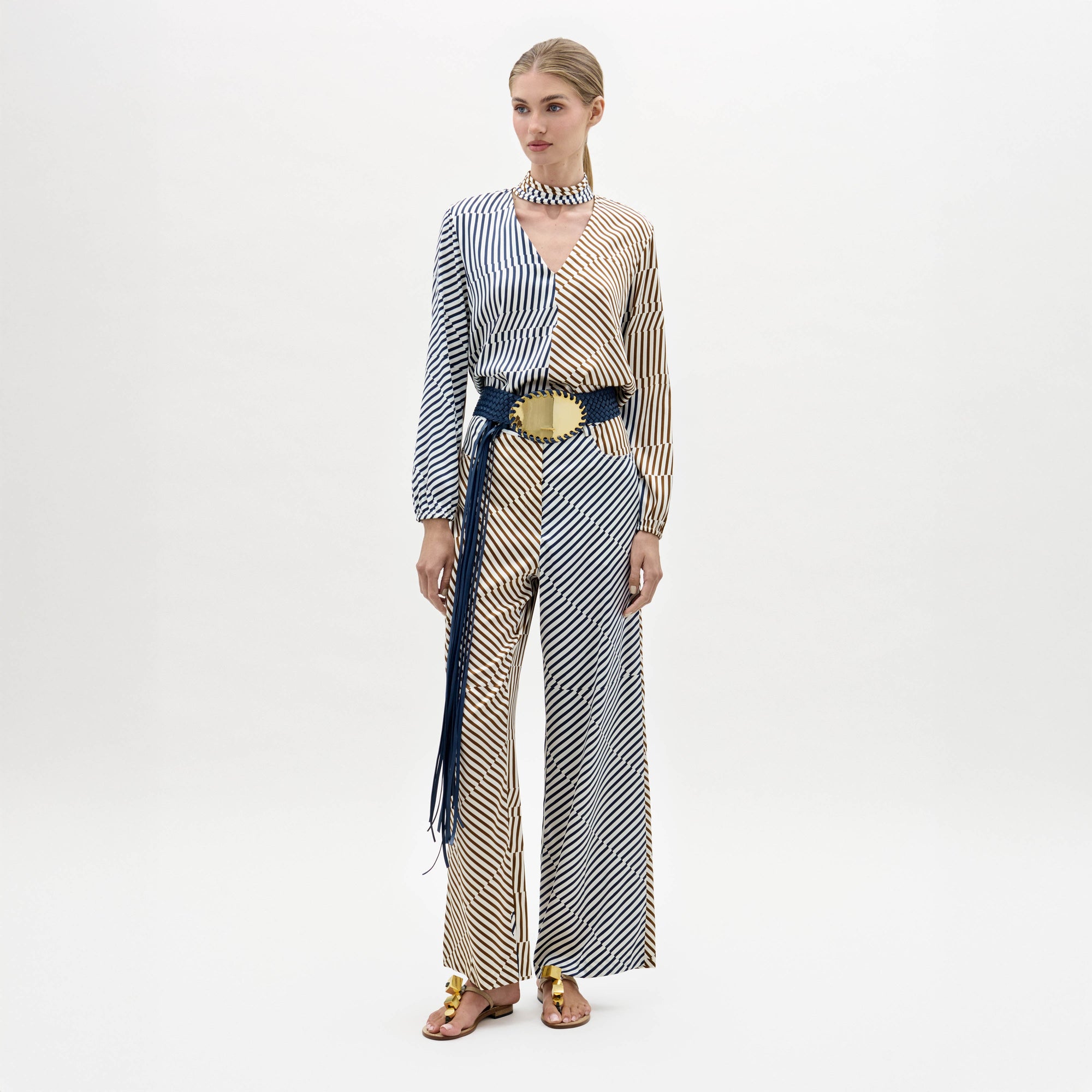 A woman stands against a plain background wearing the Emine Pant, styled with gold sandals and a matching choker.