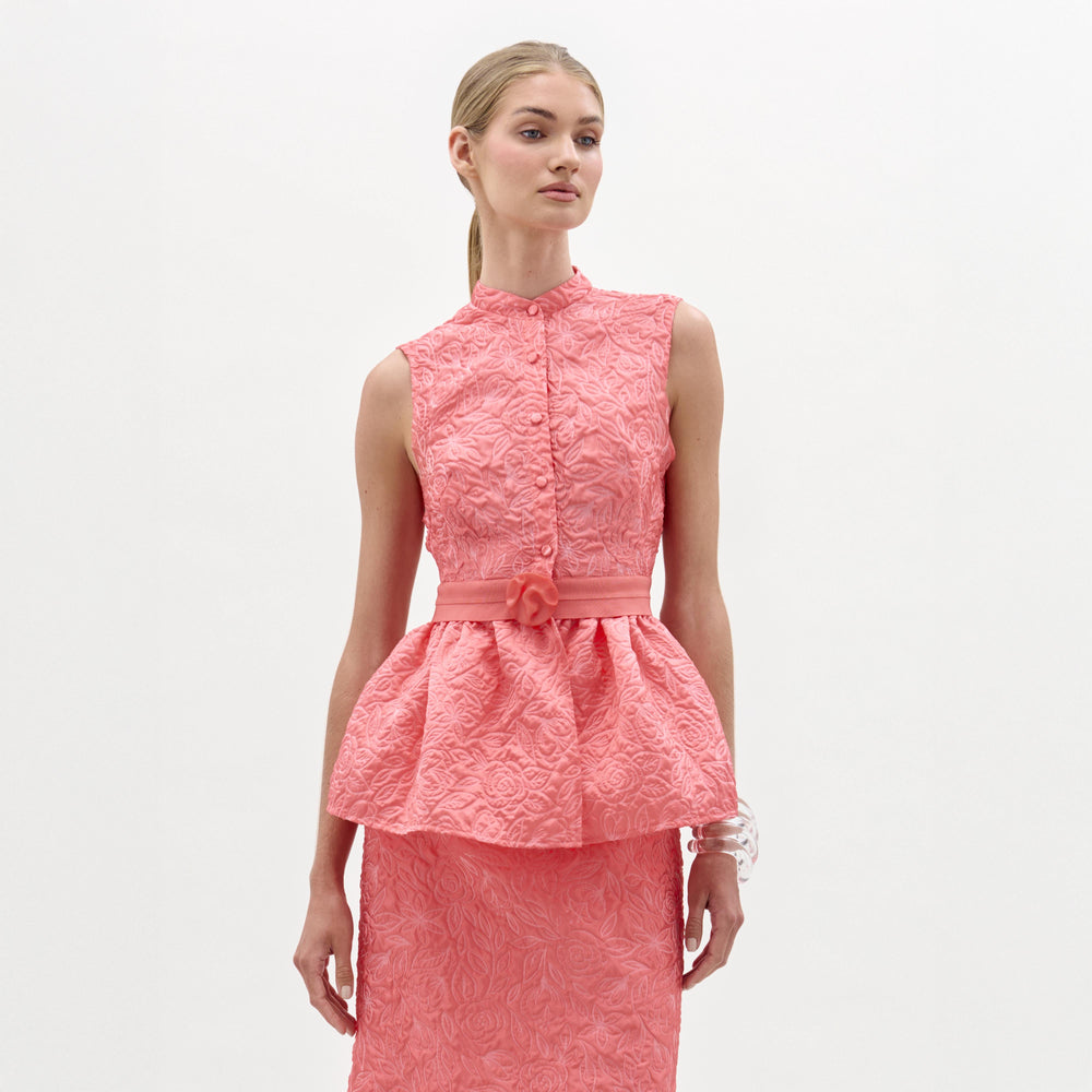 Woman wearing the Colony Blouse, a sleeveless, textured pink top with a fitted bodice and peplum hem, standing against a plain white background.