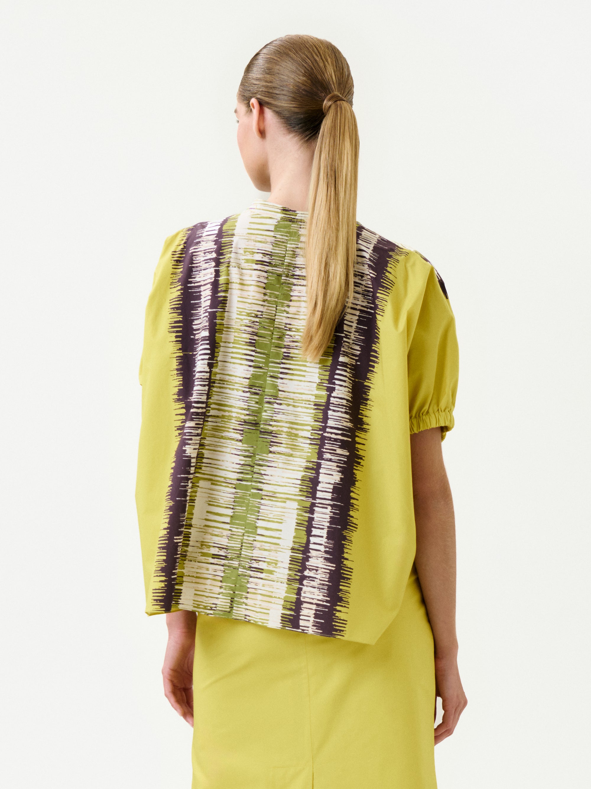 A woman with straight, light brown hair in a low ponytail faces away, wearing the Susane Blouse—a yellow top with short sleeves. Its back is made from Italian fabric featuring brown, white, and green vertical stripes.