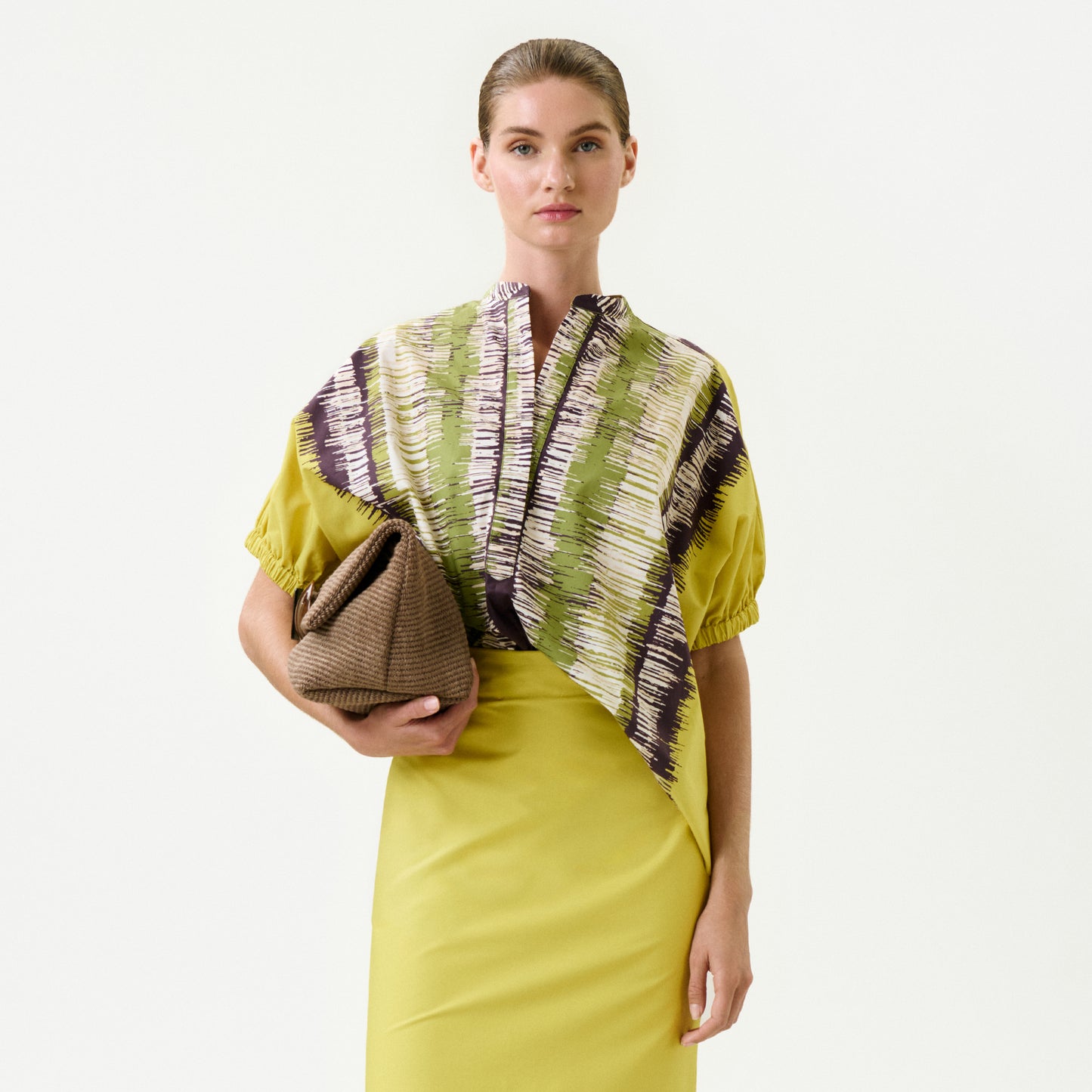 A woman wearing the Susane Blouse—a yellow dress with a patterned green, white, and purple top made from 100% cotton—holds a textured brown clutch against a plain white background.