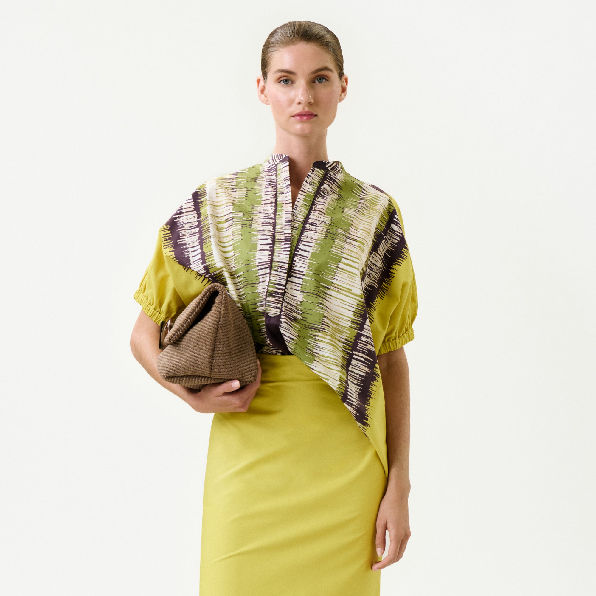 A woman wearing the Susane Blouse—a yellow dress with a patterned green, white, and purple top made from 100% cotton—holds a textured brown clutch against a plain white background.