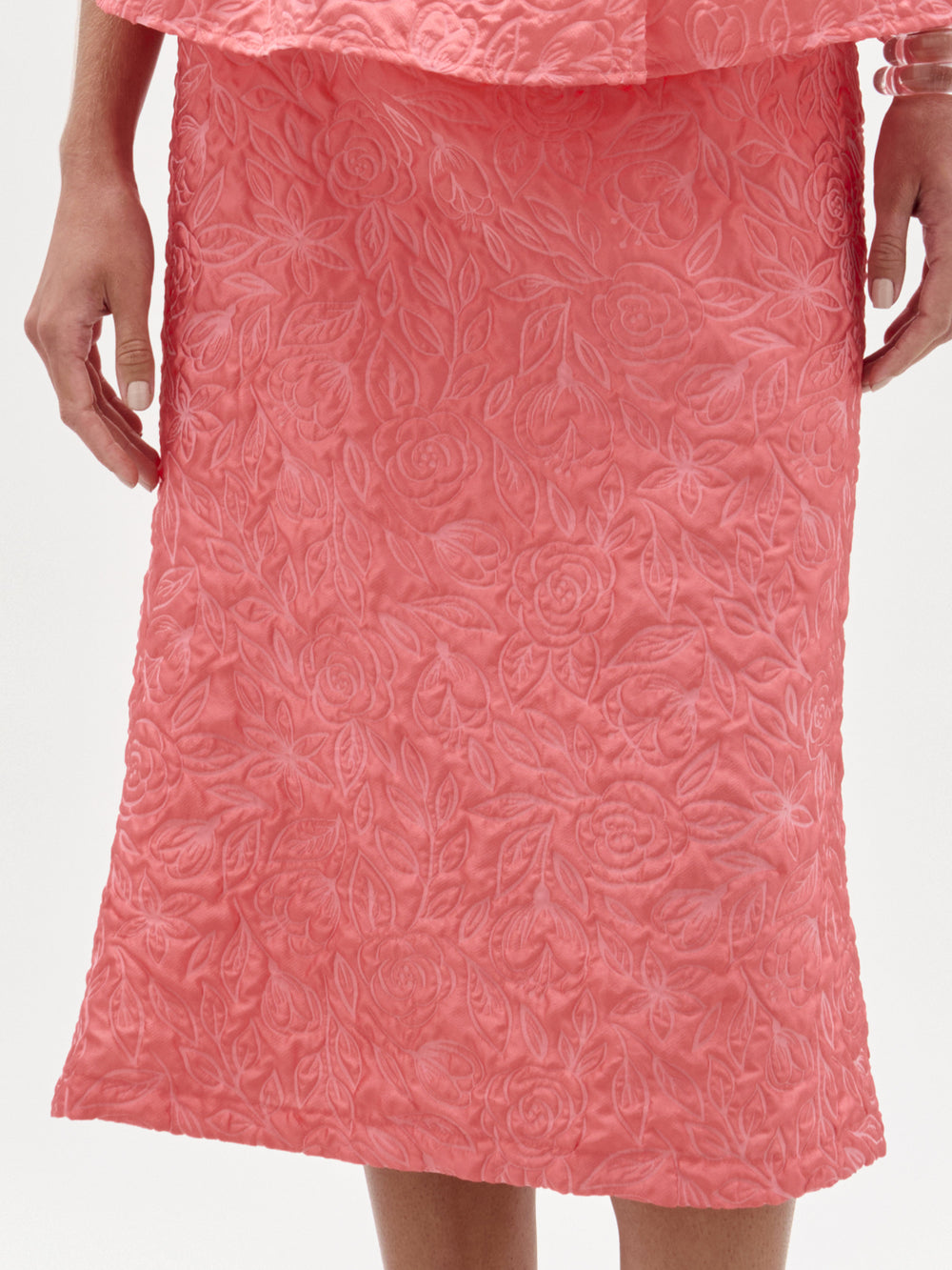 A woman stands against a white background wearing the Atira Skirt, a high-waisted midi crafted from Italian fabric, paired with a matching sleeveless pink peplum top and heeled sandals.