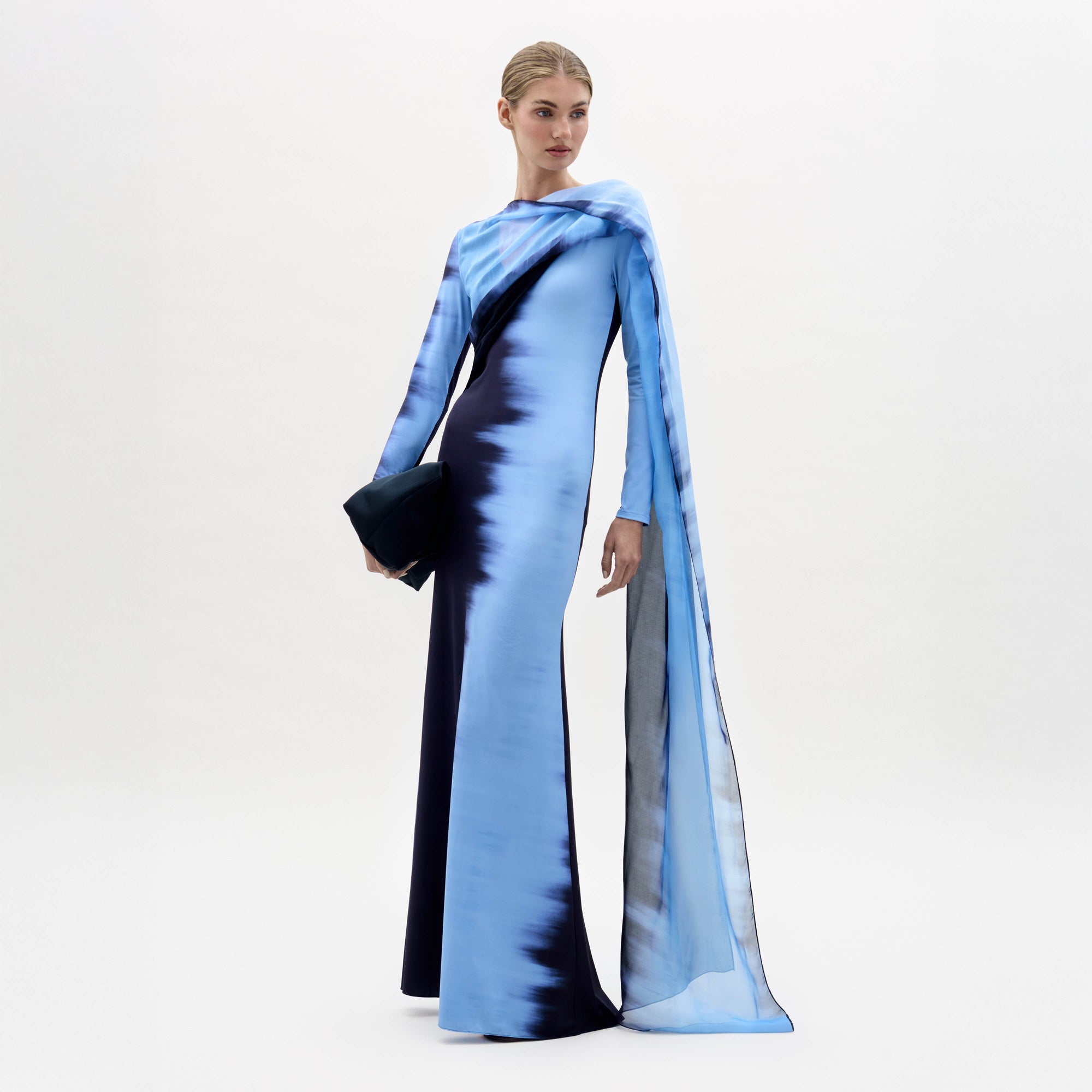 A person stands against a plain white background, wearing the Moon Dress—a long blue and black gradient gown with an attached flowing scarf—and holding a dark clutch.