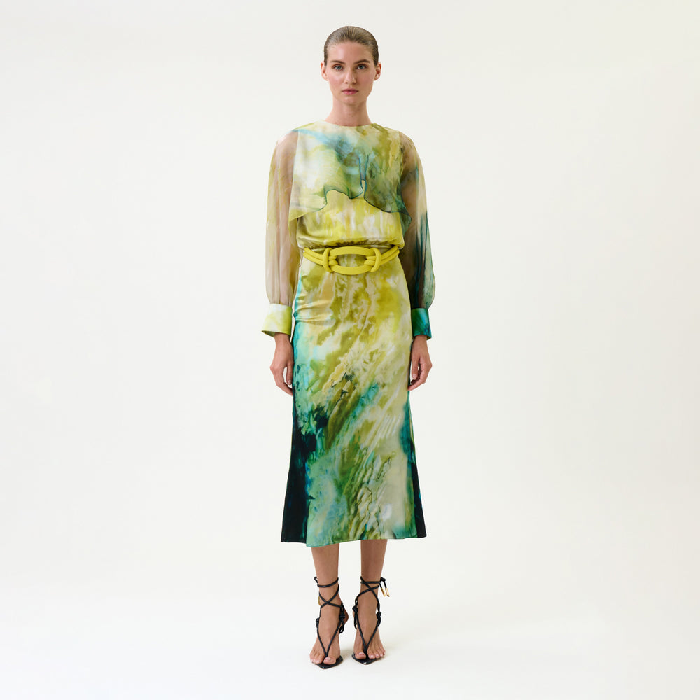 A woman wears the Rubina Dress, a long-sleeved, watercolor-patterned piece in green, yellow, and blue tones, belted at the waist. She pairs it with strappy black heels and stands against a plain white background.