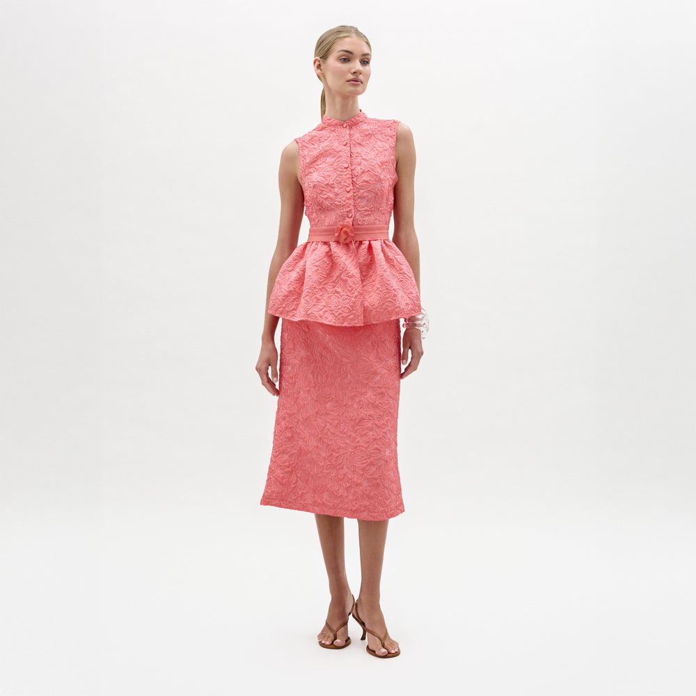 A woman stands against a white background wearing the Atira Skirt, a high-waisted midi crafted from Italian fabric, paired with a matching sleeveless pink peplum top and heeled sandals.