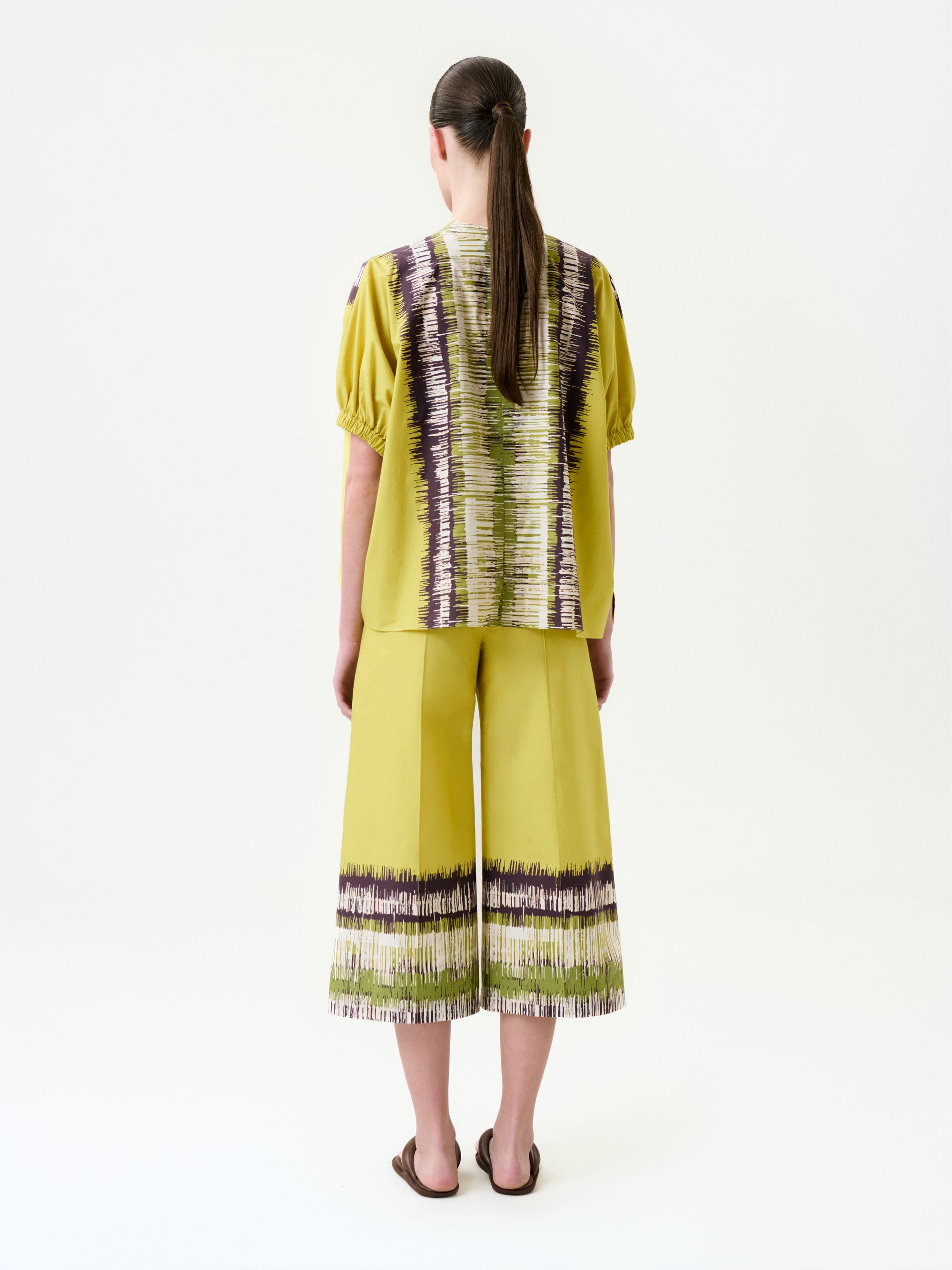 A person with long brown hair, wearing a yellow short-sleeved top and the Ilma Pant—wide-leg pants with purple and white abstract stripes in Italian fabric—stands facing away against a plain white background.