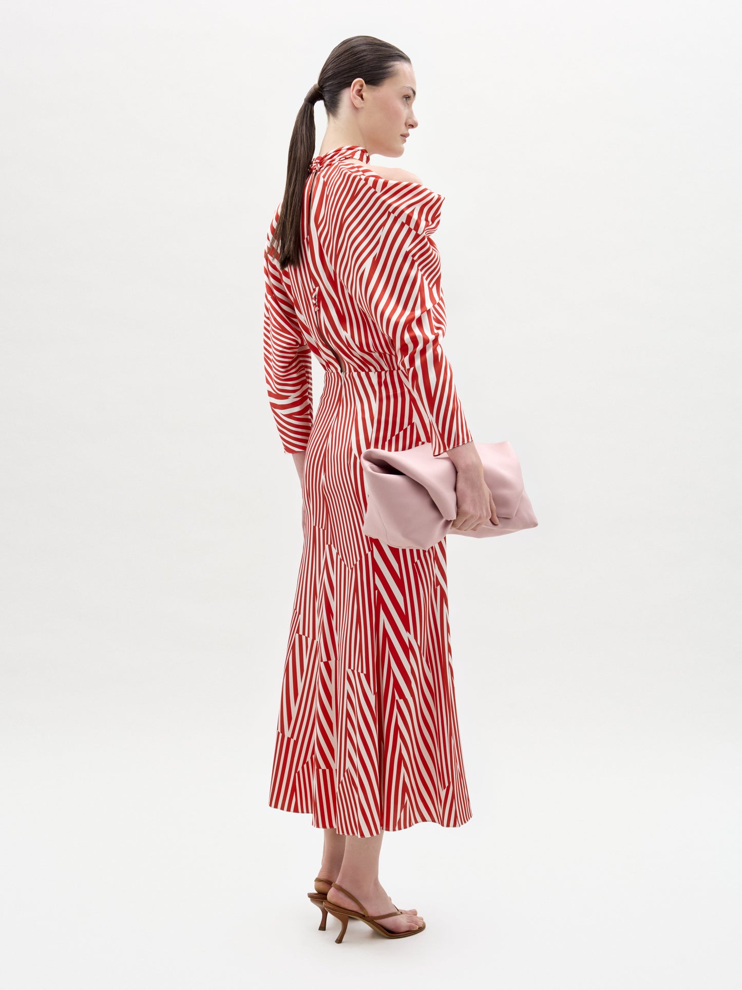 A woman stands against a plain background wearing the Lisa Dress—a red and white striped long-sleeve midi with an asymmetrical draped bodice—paired with a pink clutch and brown heeled sandals.