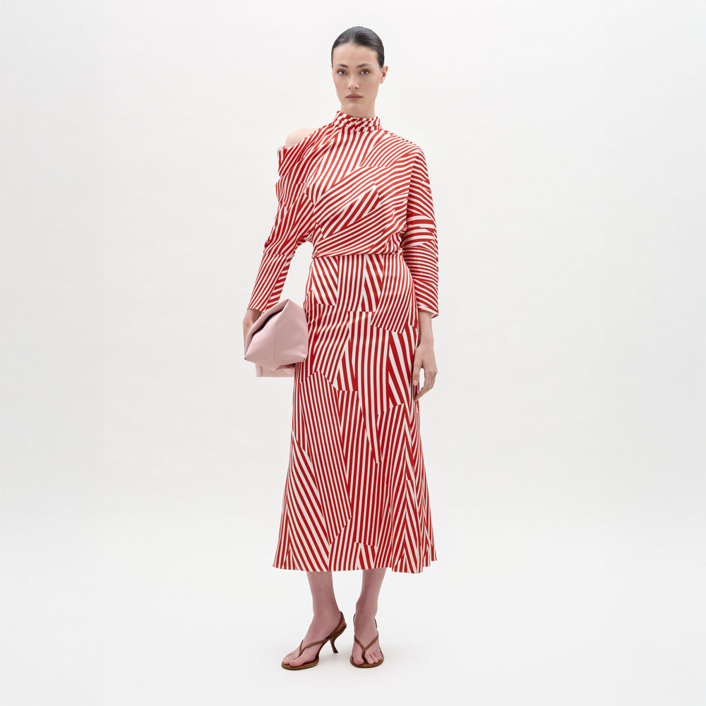 A woman stands against a plain background wearing the Lisa Dress—a red and white striped long-sleeve midi with an asymmetrical draped bodice—paired with a pink clutch and brown heeled sandals.