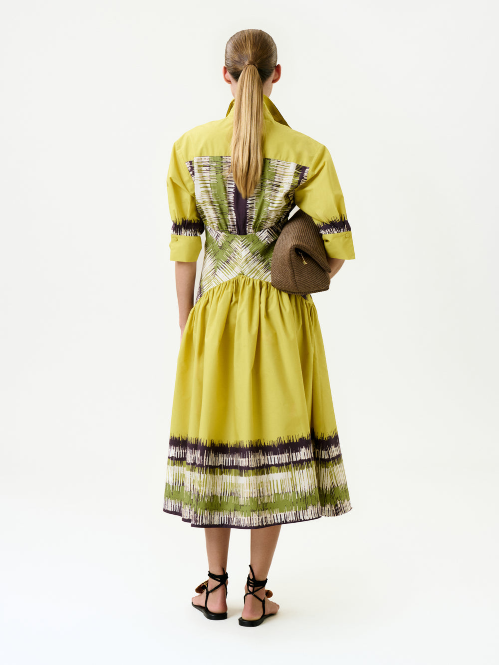 A woman with long, straight hair in a low ponytail is seen from behind against a plain white background, wearing the yellow Indy Dress crafted from Italian fabric with patterned accents, black sandals, and holding a brown textured clutch.