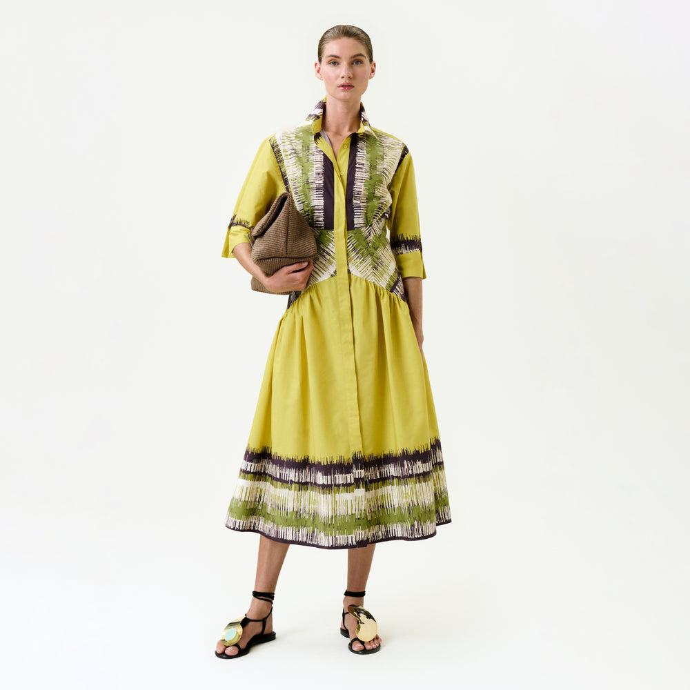 A woman stands against a plain background, wearing the Indy Dress in yellow with green and purple patterns made from Italian fabric, paired with black-and-beige sandals and holding a brown textured clutch under her arm.