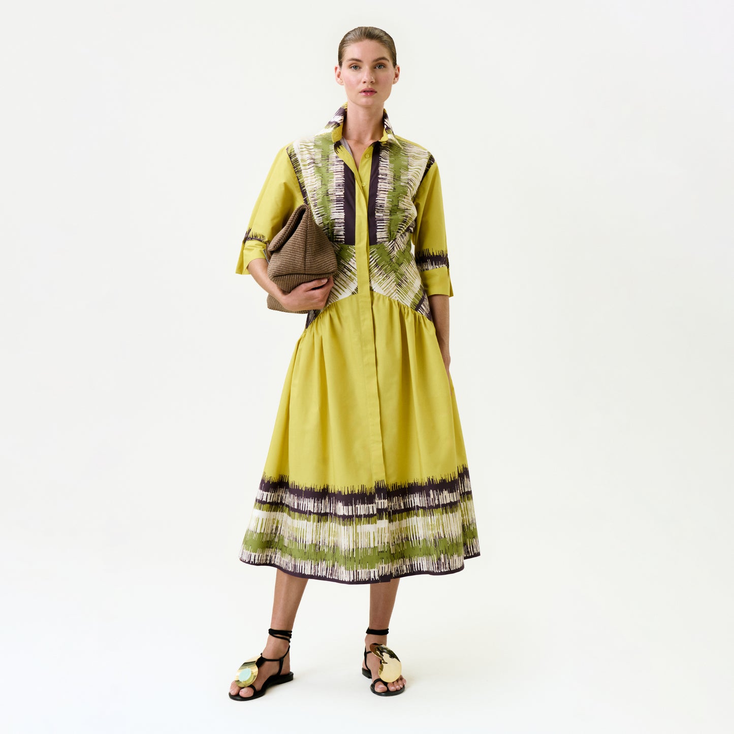 A woman stands against a plain background, wearing the Indy Dress in yellow with green and purple patterns made from Italian fabric, paired with black-and-beige sandals and holding a brown textured clutch under her arm.