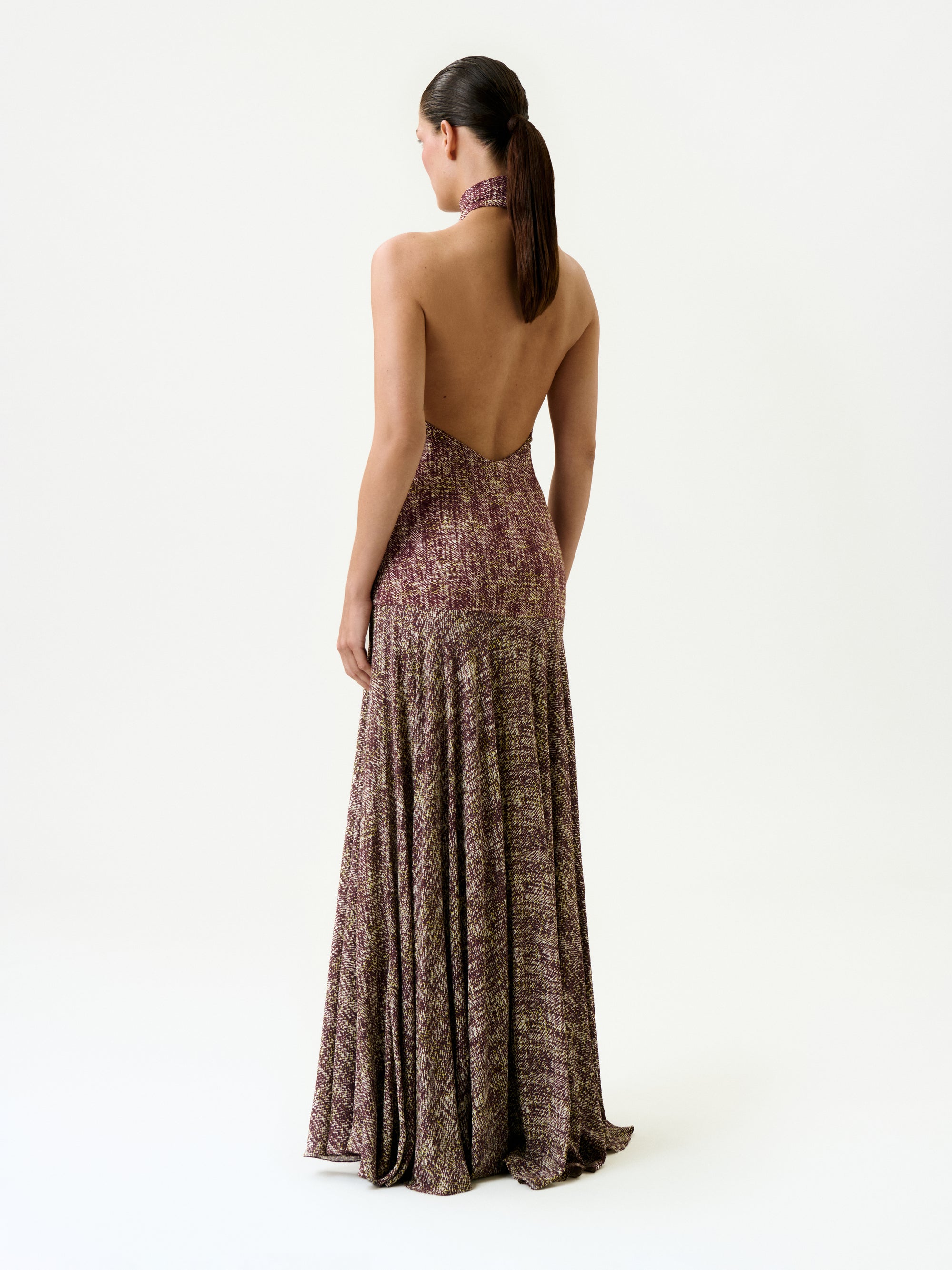 A woman with dark hair in a ponytail faces away from the camera, wearing the Leonore Dress—a halter-neck, backless maxi dress featuring a multicolored textured pattern, made in Colombia—against a plain white background.