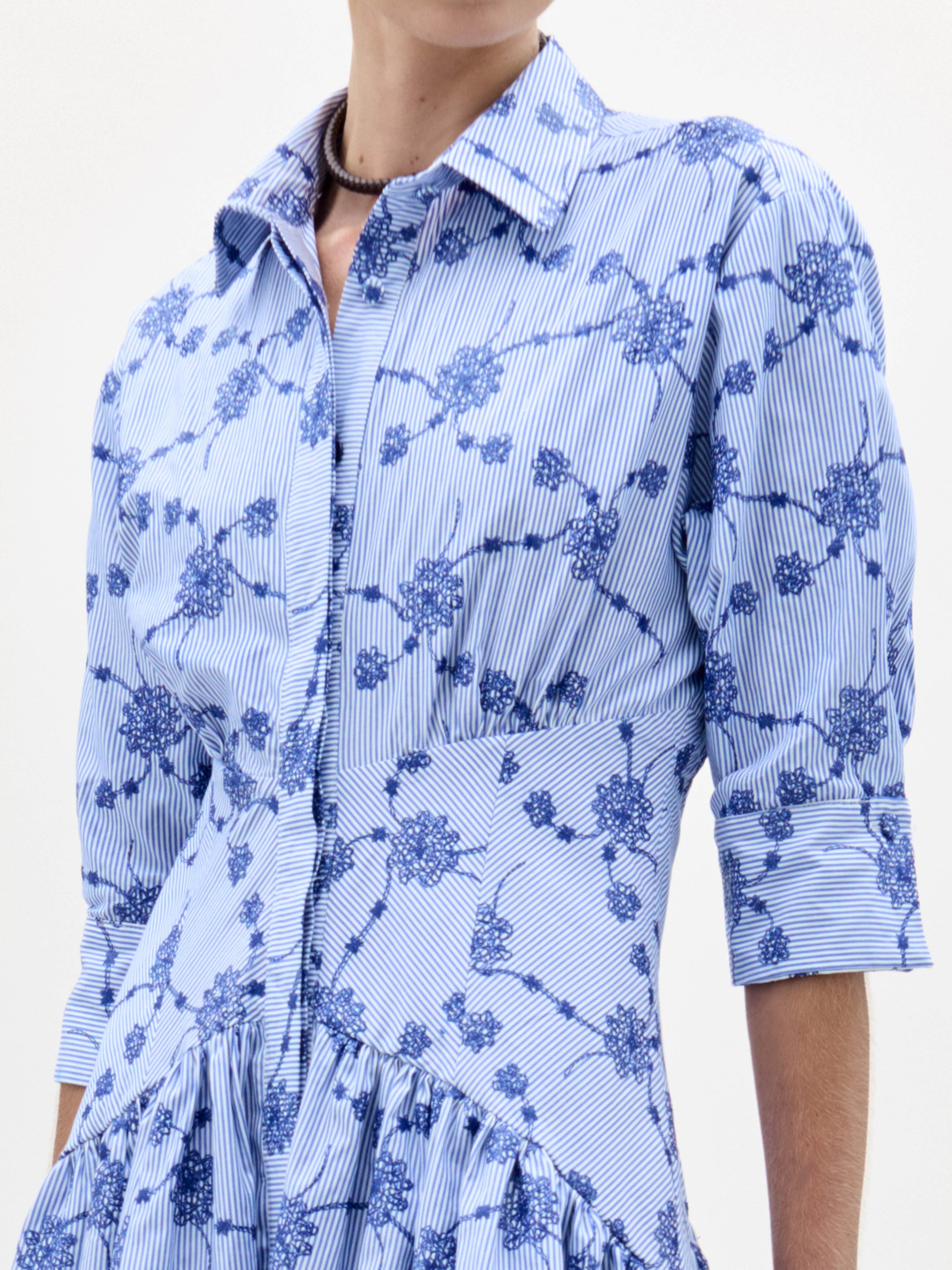 A woman stands against a plain white background wearing the Indy Dress—a light blue, patterned, collared midi dress with three-quarter sleeves—paired with brown sandals and a fringed handbag.