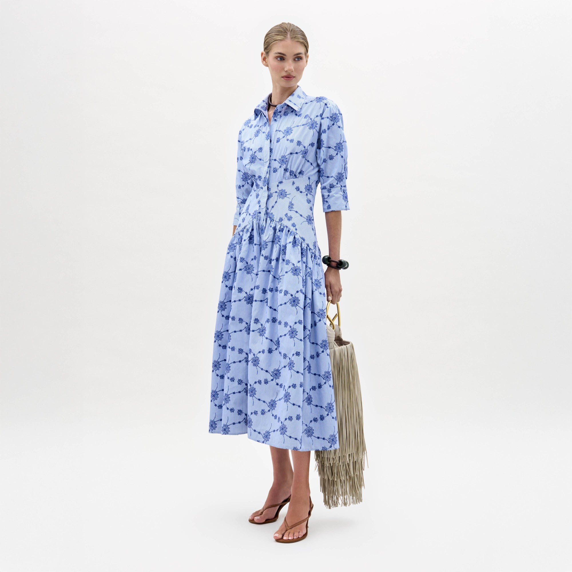 A woman stands against a plain white background wearing the Indy Dress—a light blue, patterned, collared midi dress with three-quarter sleeves—paired with brown sandals and a fringed handbag.