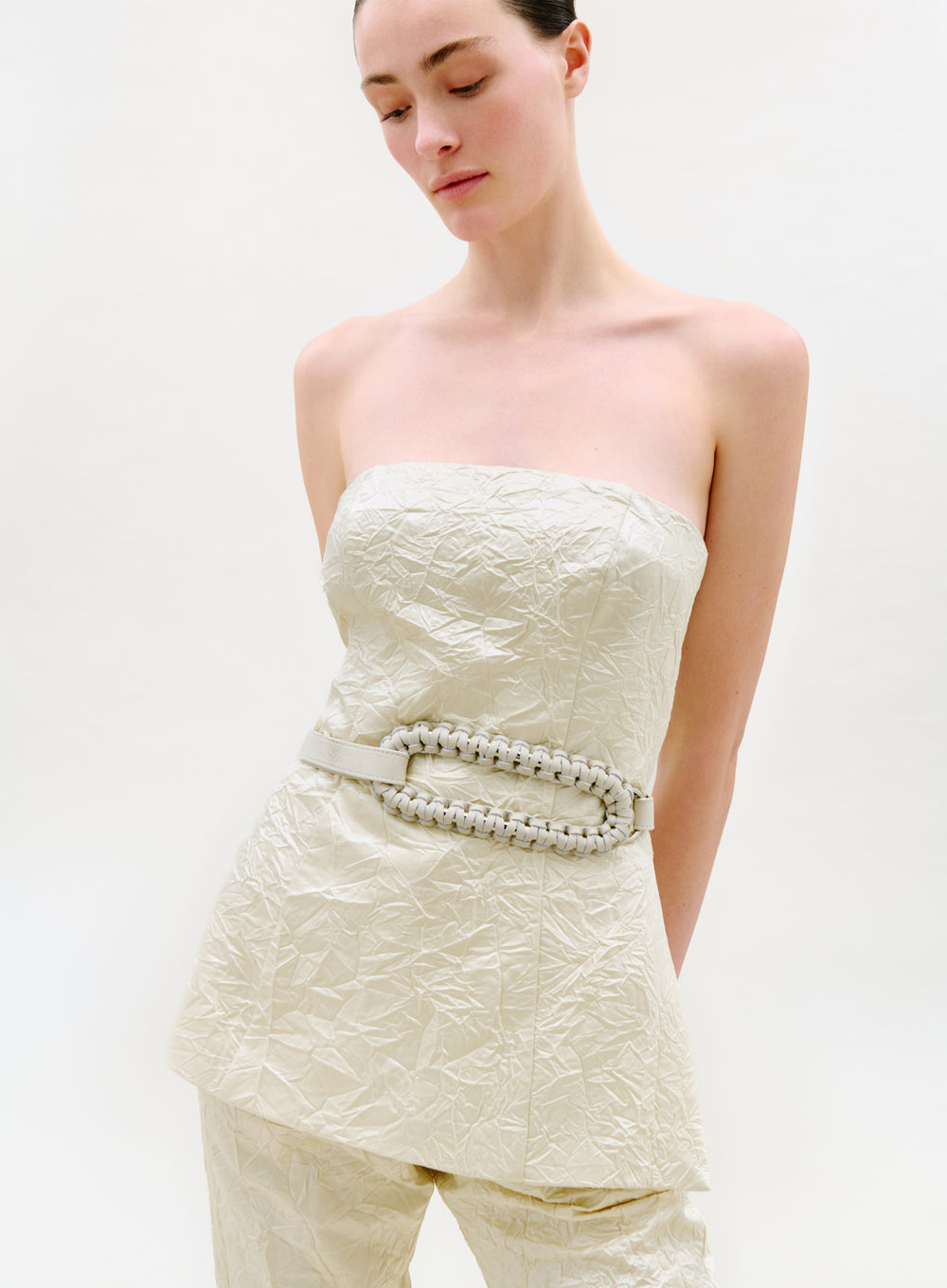 A woman stands against a plain light background, looking downward with a serene expression. She wears the Astor Top—strapless and textured in cream—with matching pants and a wide, braided belt.