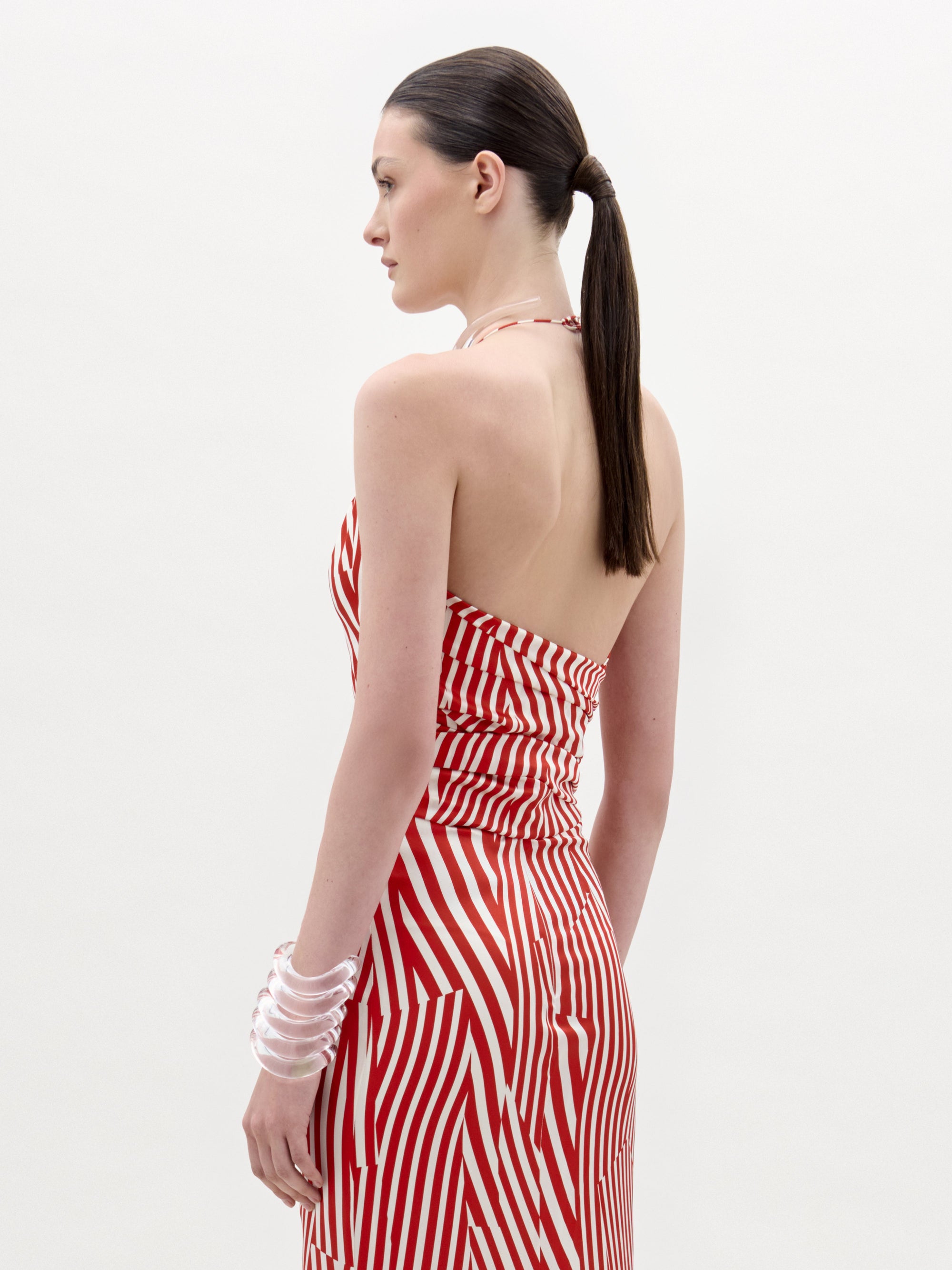 A woman stands against a plain background wearing the Carmela Blouse, which features sleeveless red and white stripes with geometric patterns and a halter neckline, accessorized with a clear chunky bracelet.