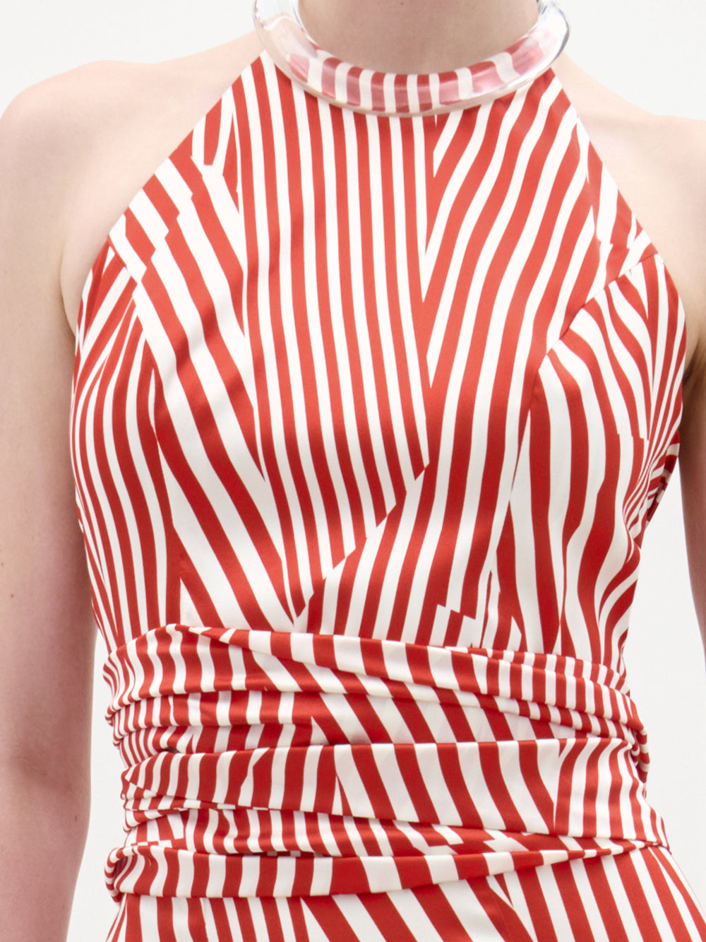 A woman stands against a plain background wearing the Carmela Blouse, which features sleeveless red and white stripes with geometric patterns and a halter neckline, accessorized with a clear chunky bracelet.
