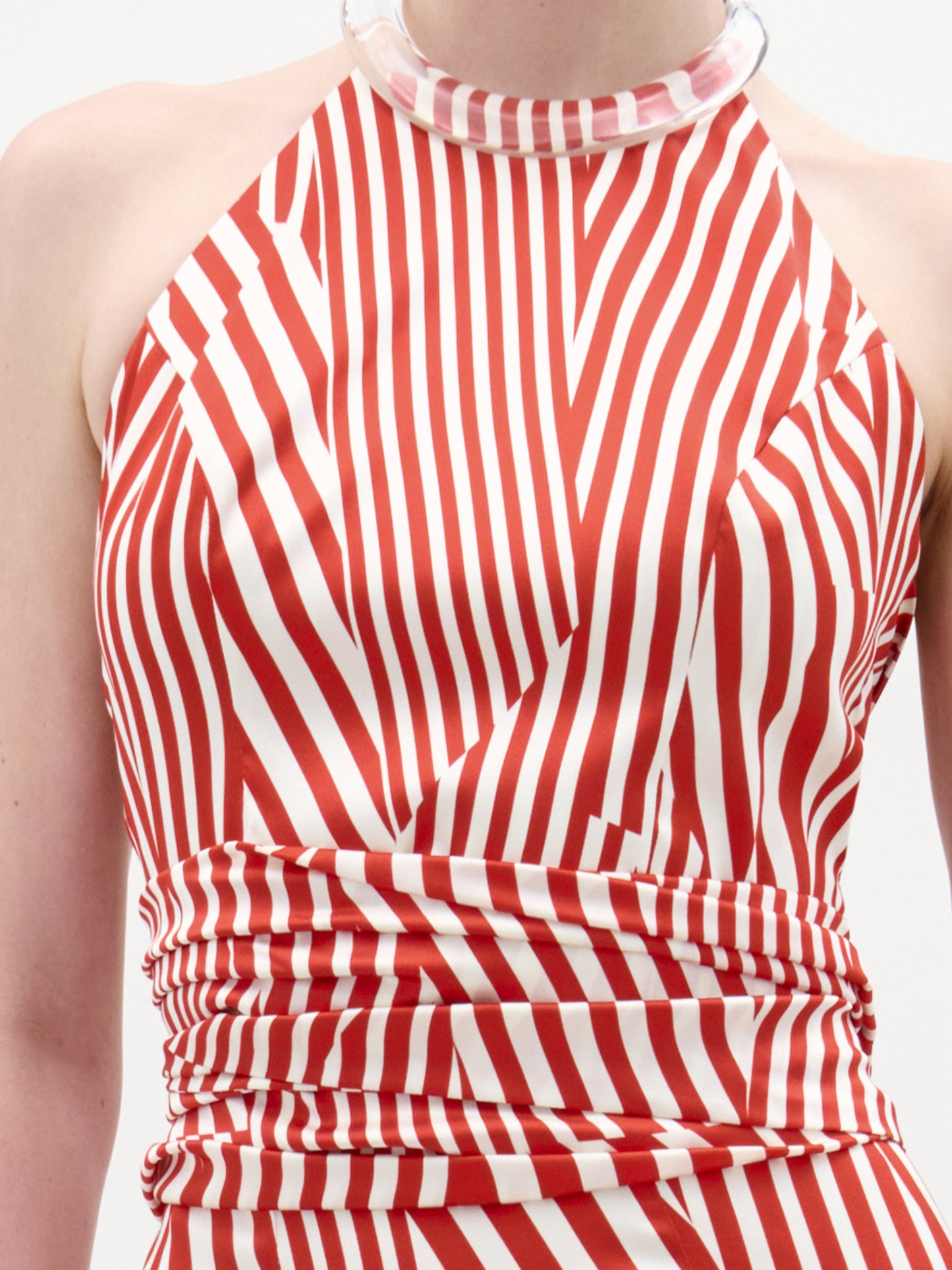 A woman stands against a plain background wearing the Carmela Blouse, which features sleeveless red and white stripes with geometric patterns and a halter neckline, accessorized with a clear chunky bracelet.