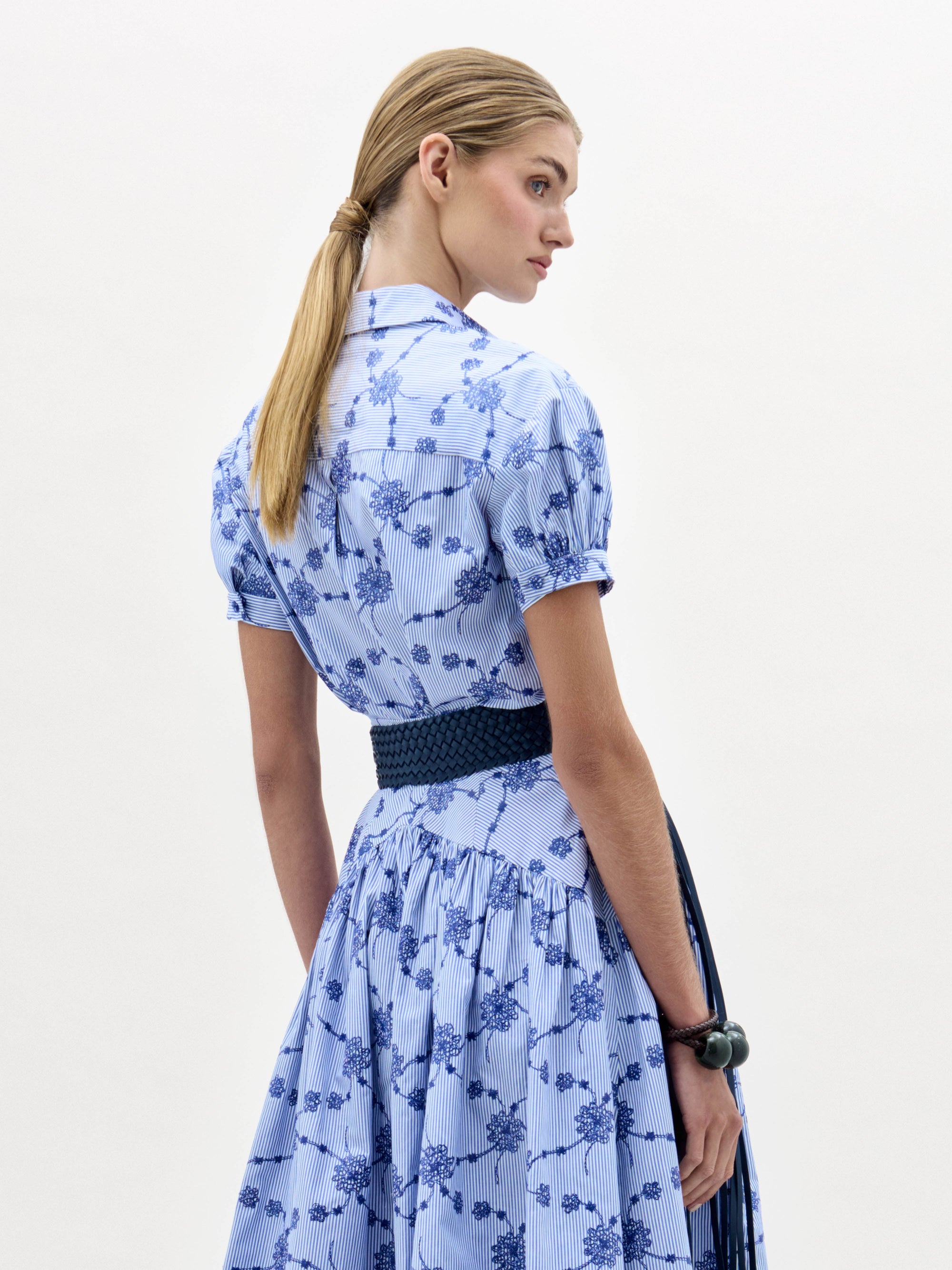 A woman in the Ileana Blouse, a light blue short-sleeved, collared top with a blue floral pattern and a wide dark blue belt with a large gold buckle, stands against a plain light background.