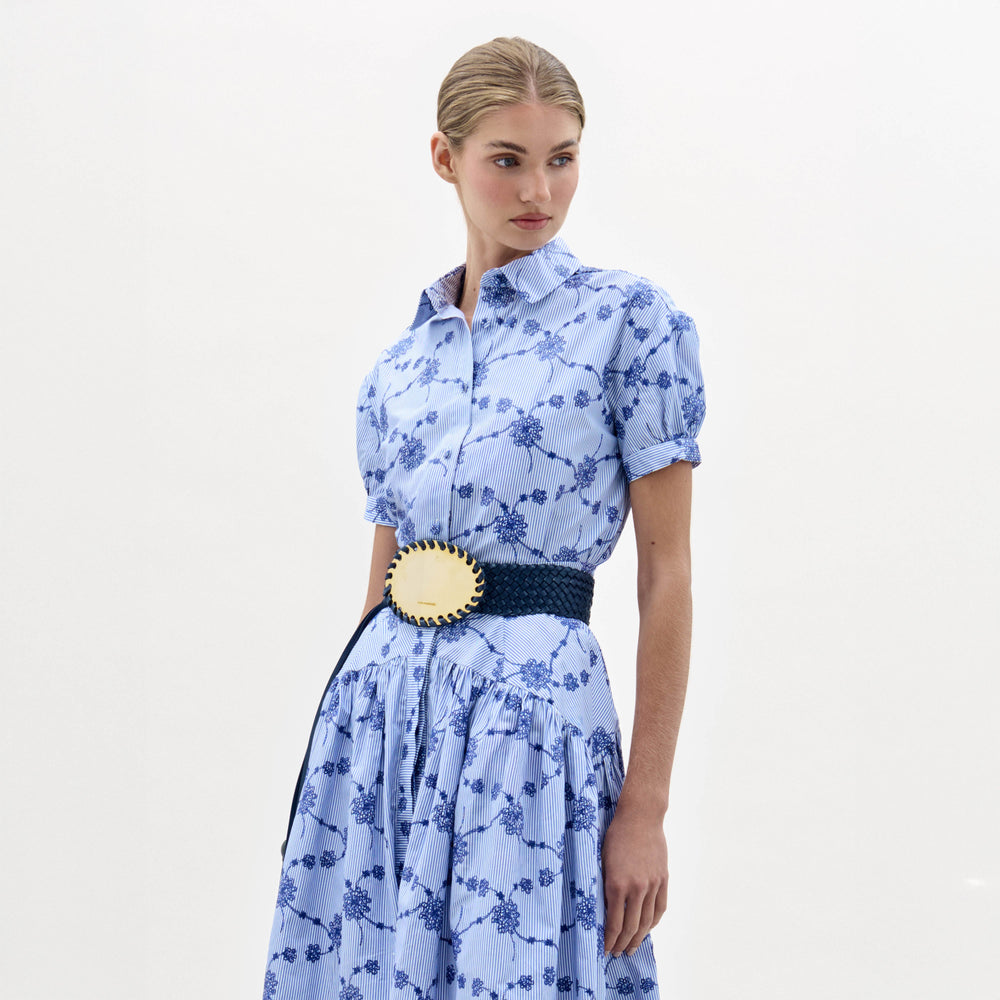 A woman in the Ileana Blouse, a light blue short-sleeved, collared top with a blue floral pattern and a wide dark blue belt with a large gold buckle, stands against a plain light background.