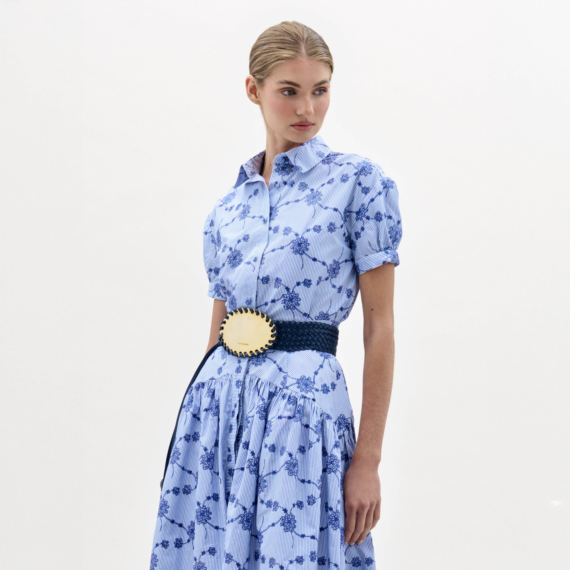 A woman in the Ileana Blouse, a light blue short-sleeved, collared top with a blue floral pattern and a wide dark blue belt with a large gold buckle, stands against a plain light background.