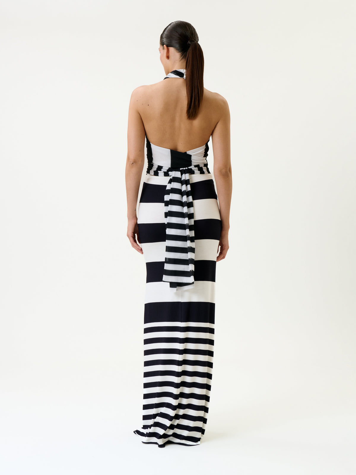 A woman stands facing away, wearing the Tove Dress—a long, sleeveless black and white striped dress with a halter neck and a bow tied at the back. Her hair is pulled back in a low ponytail, against a plain white background.