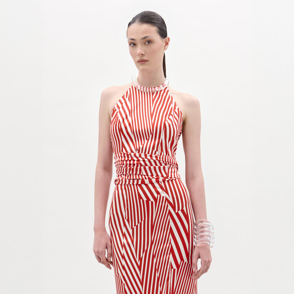 A woman stands against a plain background wearing the Carmela Blouse, which features sleeveless red and white stripes with geometric patterns and a halter neckline, accessorized with a clear chunky bracelet.
