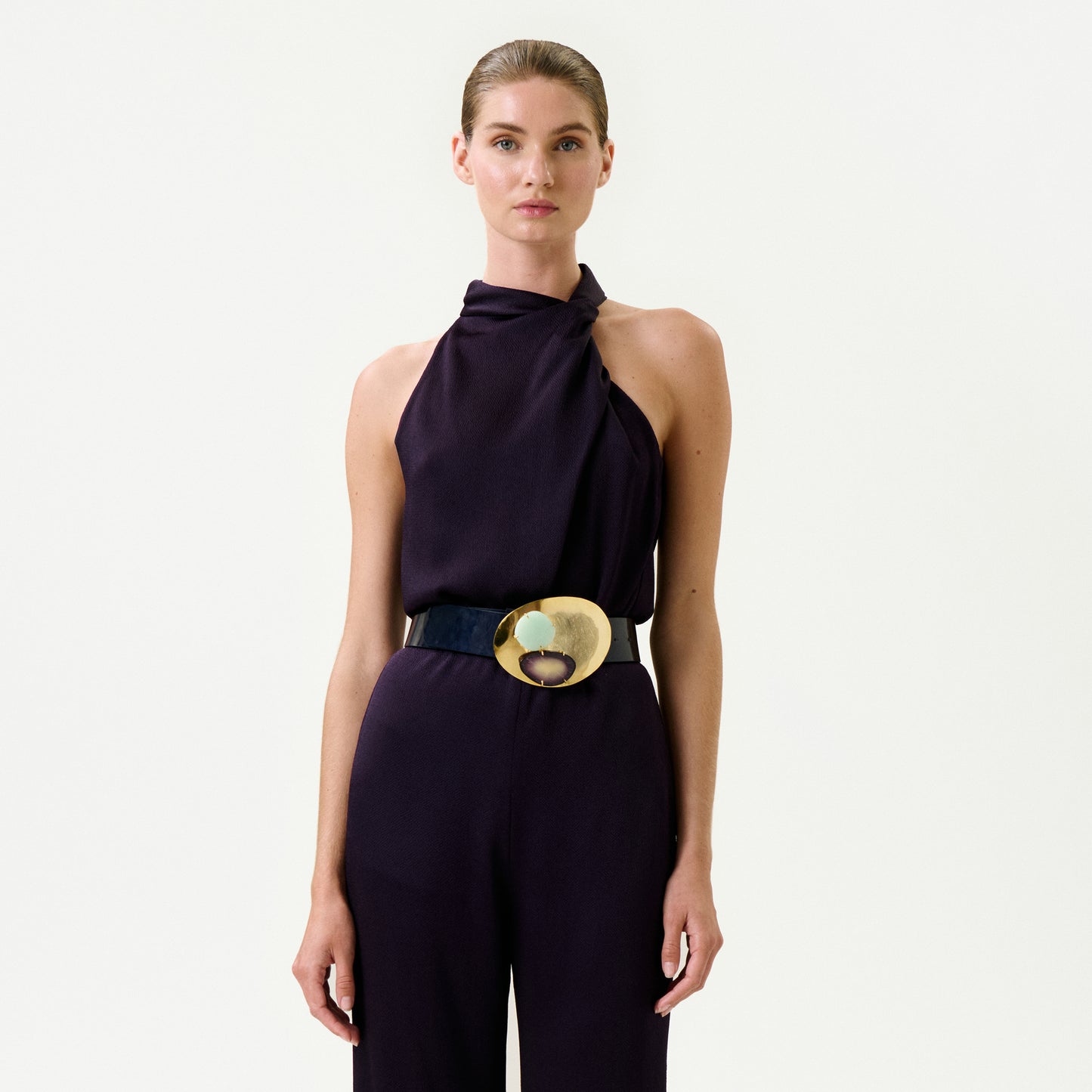 A woman stands against a white background wearing the Naili Blouse—a sleeveless, dark purple Italian-fabric top with a high neckline—paired with a wide black belt with a large, round gold buckle. Her hair is pulled back and she has a neutral expression.
