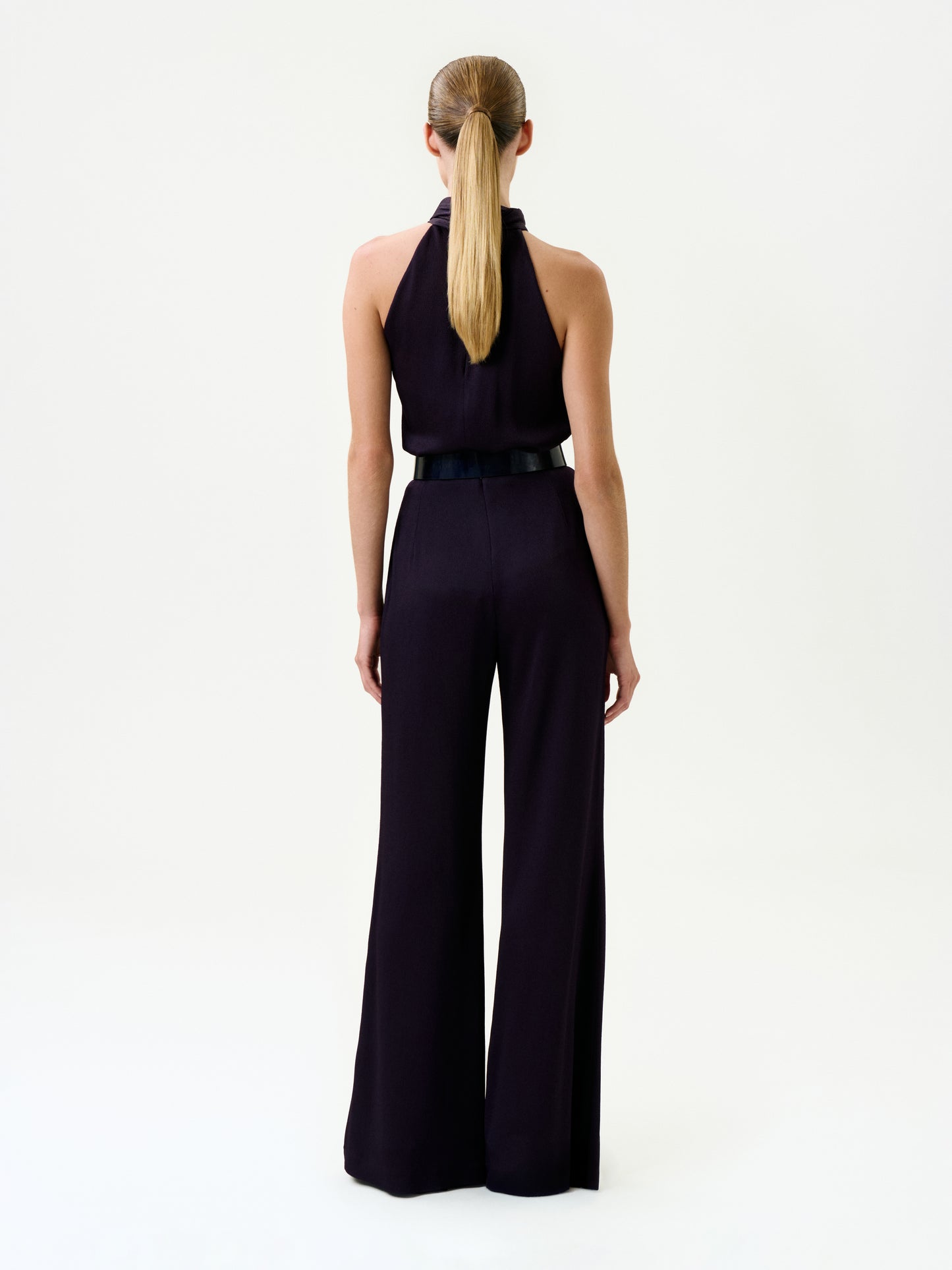A person with long, straight hair in a low ponytail is seen from behind, wearing the Palermo Pant—a sleeveless black halter-neck jumpsuit with wide-leg pants and a black belt, made from luxurious Italian fabric—set against a plain white background.