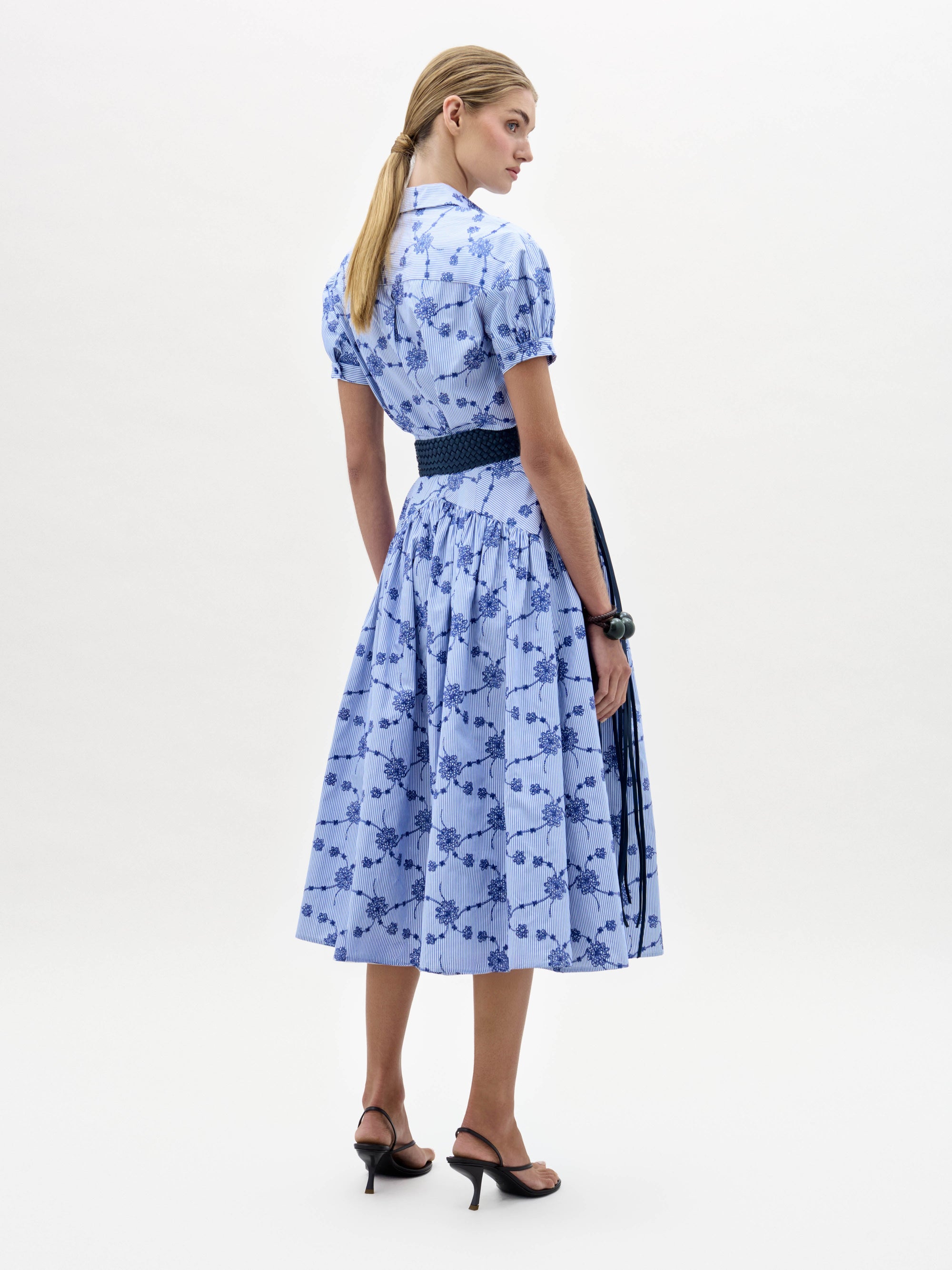 A woman wearing the Narel Skirt, styled with a light blue short-sleeve floral-print top, a wide dark belt, and black heeled sandals, stands against a plain white background.