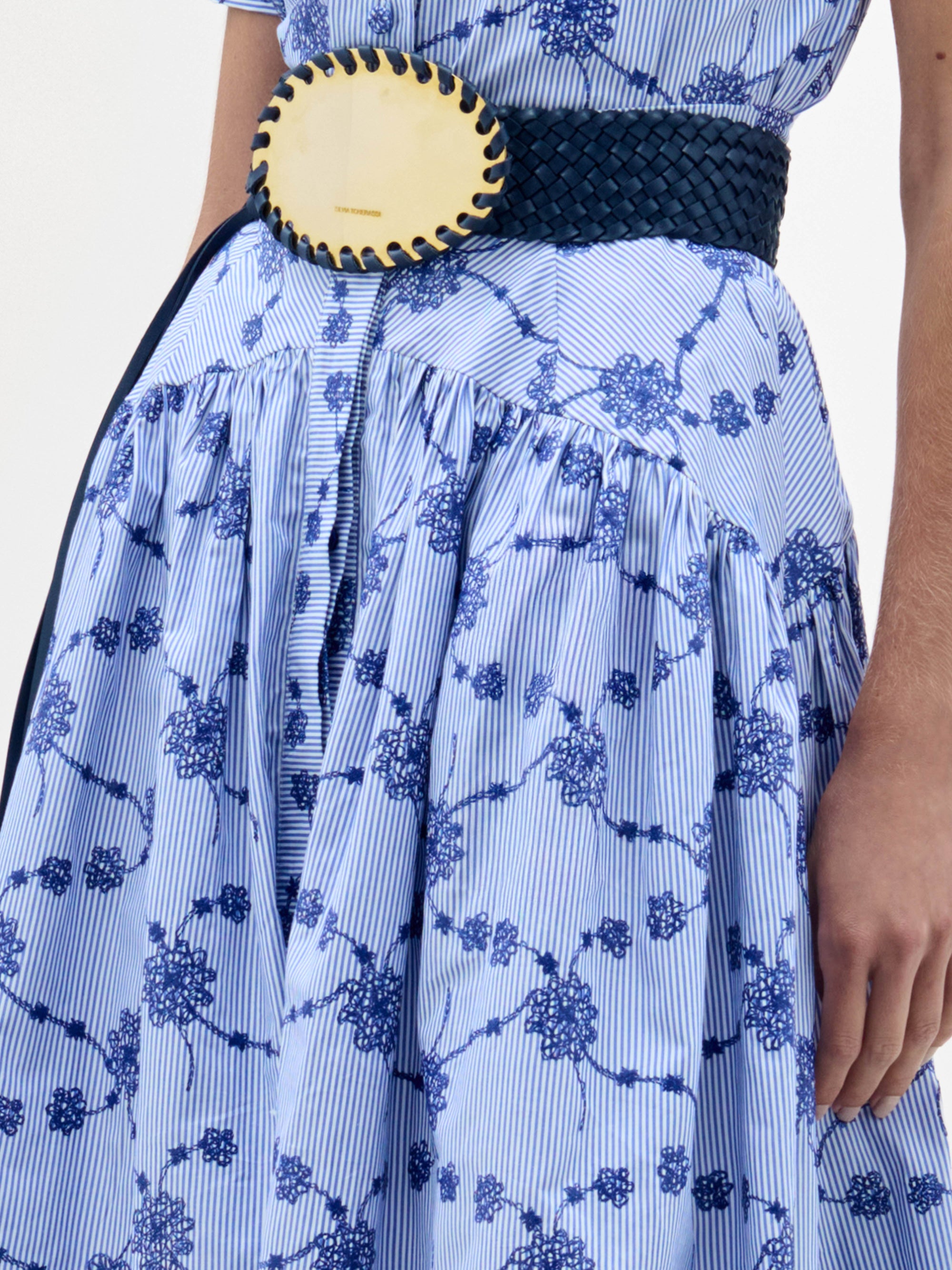 A woman wearing the Narel Skirt, styled with a light blue short-sleeve floral-print top, a wide dark belt, and black heeled sandals, stands against a plain white background.