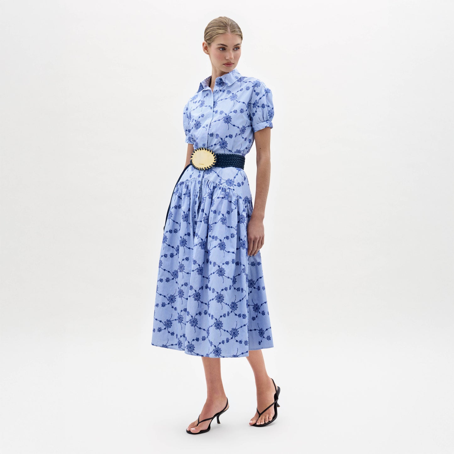 A woman wearing the Narel Skirt, styled with a light blue short-sleeve floral-print top, a wide dark belt, and black heeled sandals, stands against a plain white background.