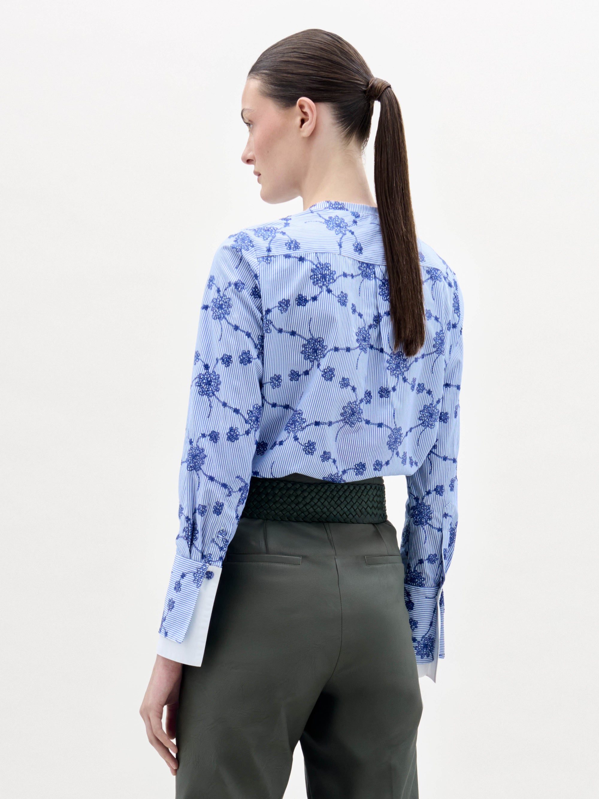 A woman stands against a plain background wearing the Cubillos Blouse, a blue floral top, paired with green high-waisted pants and a wide green belt featuring a large gold buckle and long fringe.