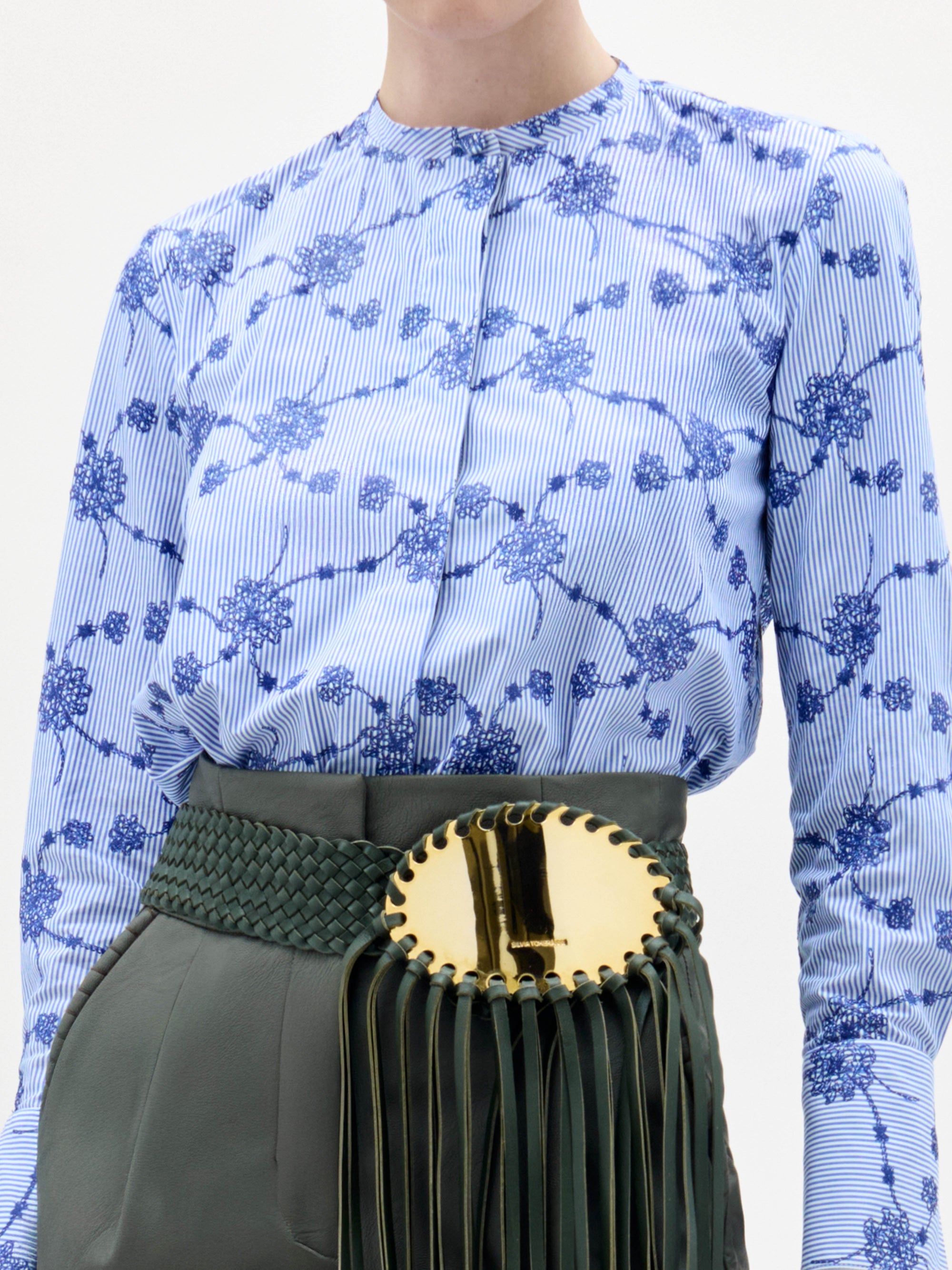 A woman stands against a plain background wearing the Cubillos Blouse, a blue floral top, paired with green high-waisted pants and a wide green belt featuring a large gold buckle and long fringe.