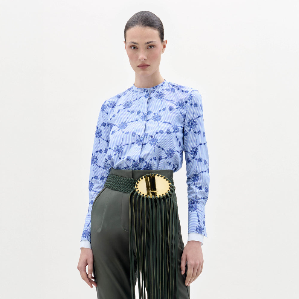 A woman stands against a plain background wearing the Cubillos Blouse, a blue floral top, paired with green high-waisted pants and a wide green belt featuring a large gold buckle and long fringe.