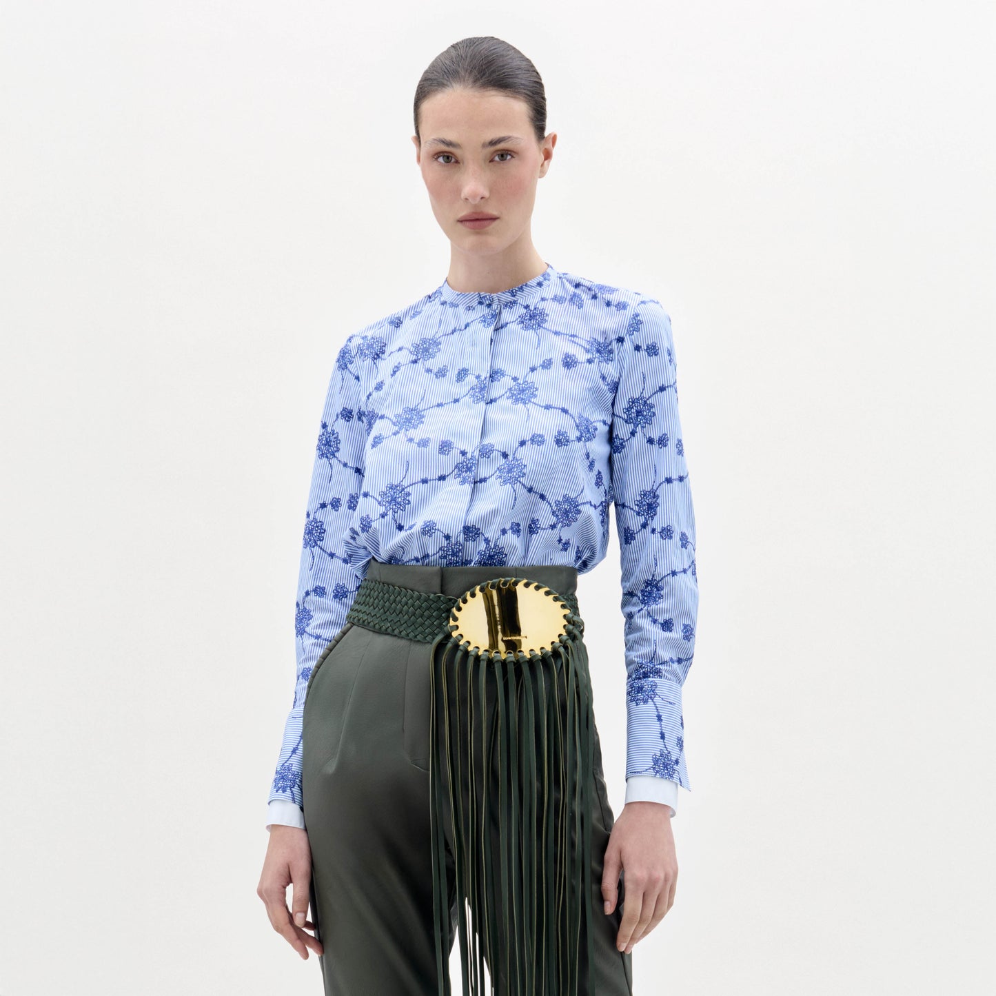 A woman stands against a plain background wearing the Cubillos Blouse, a blue floral top, paired with green high-waisted pants and a wide green belt featuring a large gold buckle and long fringe.