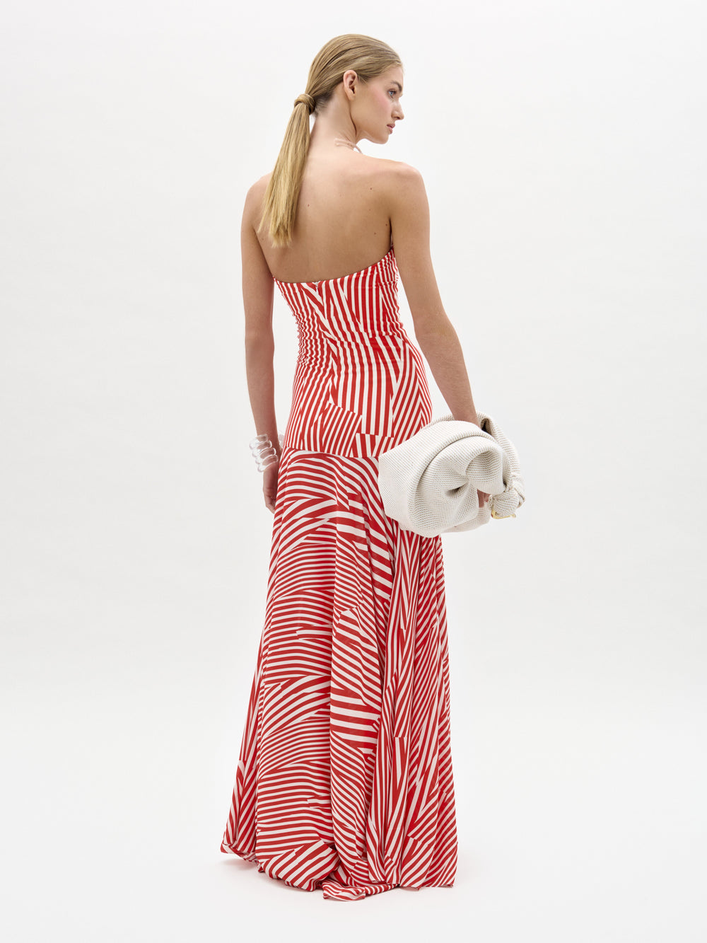 A woman stands against a plain white background in the Muriel Dress—a red and white striped, strapless maxi with a fitted bodice crafted from luxurious Italian fabric. She accessorizes with pearls, a white clutch, and slicked-back hair.