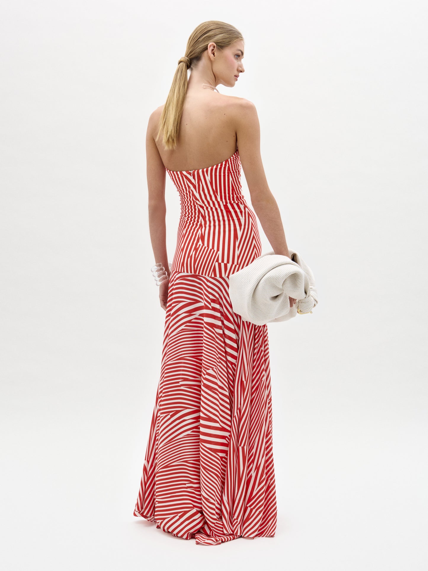 A woman stands against a plain white background in the Muriel Dress—a red and white striped, strapless maxi with a fitted bodice crafted from luxurious Italian fabric. She accessorizes with pearls, a white clutch, and slicked-back hair.