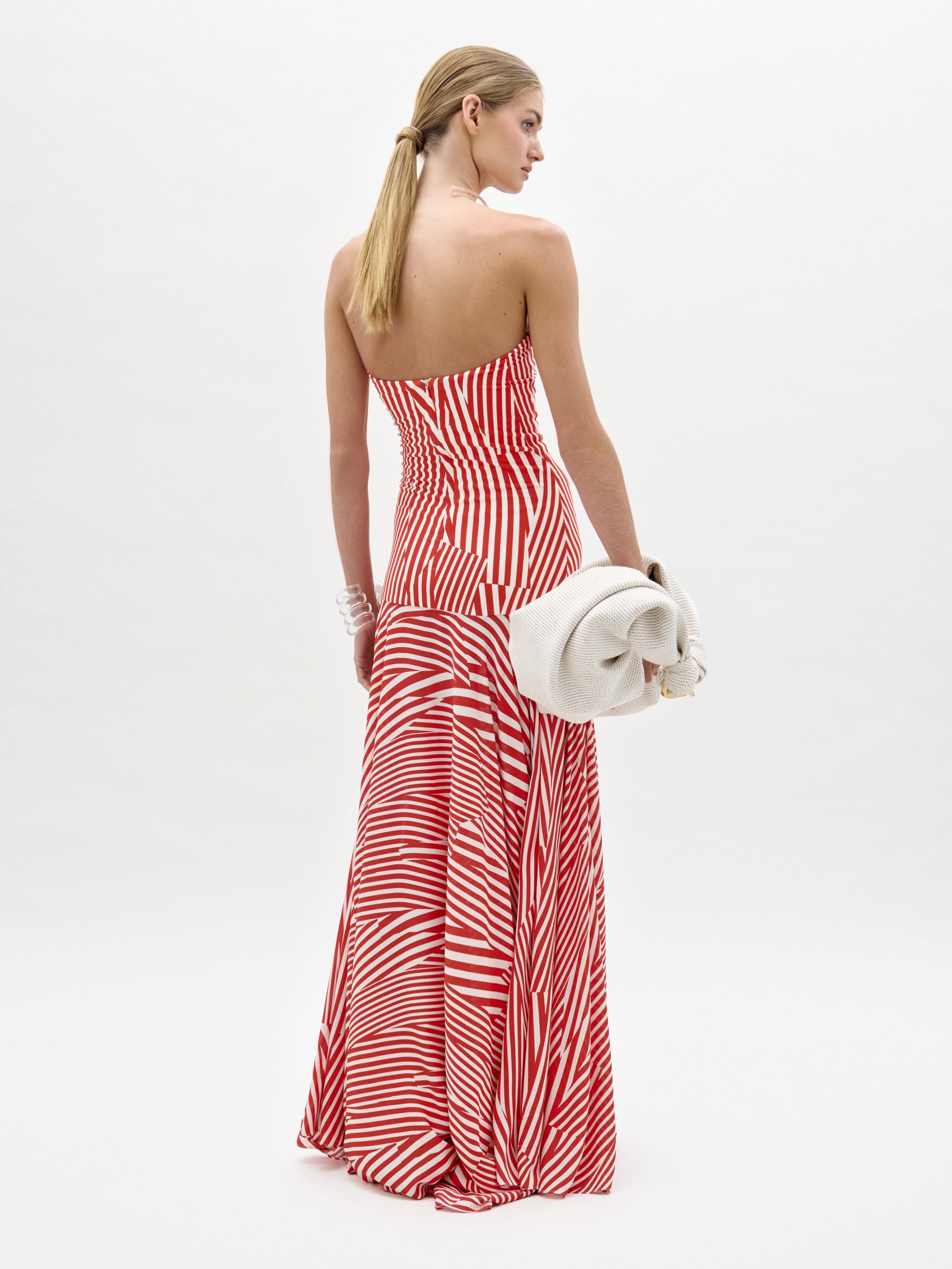 A woman stands against a plain white background in the Muriel Dress—a red and white striped, strapless maxi with a fitted bodice crafted from luxurious Italian fabric. She accessorizes with pearls, a white clutch, and slicked-back hair.