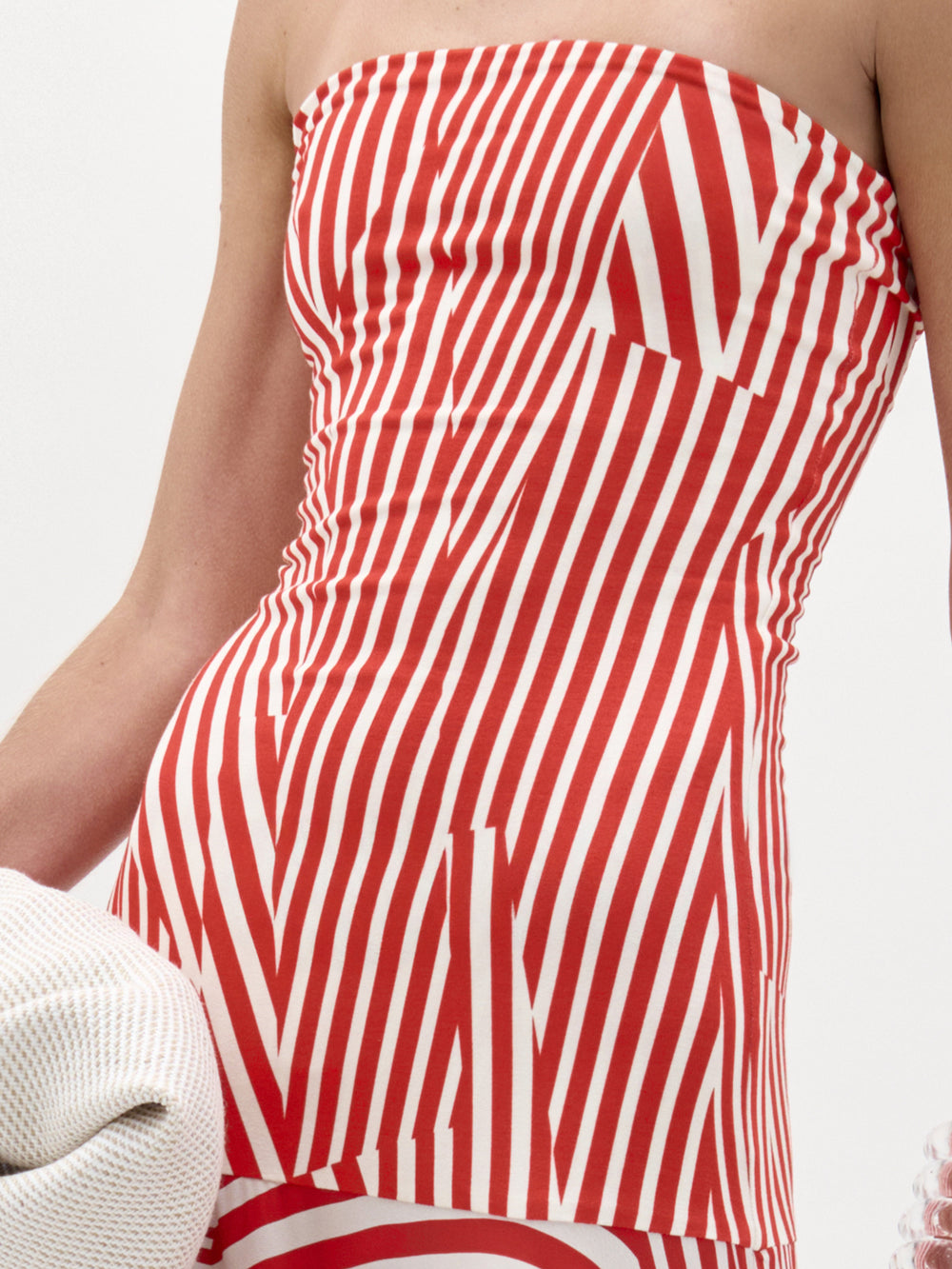 A woman stands against a plain white background in the Muriel Dress—a red and white striped, strapless maxi with a fitted bodice crafted from luxurious Italian fabric. She accessorizes with pearls, a white clutch, and slicked-back hair.