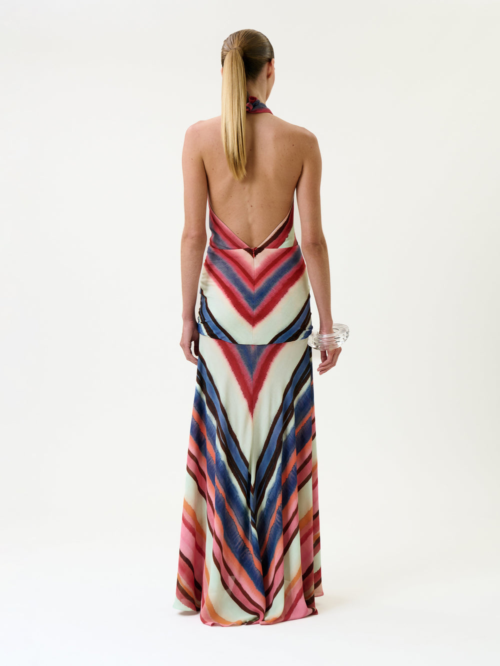 A person with long blonde hair in a ponytail wears the Patsy Dress—a backless halter design featuring colorful chevron stripes—paired with clear bangles, standing against a plain white background.