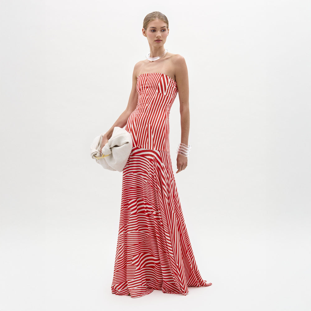 A woman stands against a plain white background in the Muriel Dress—a red and white striped, strapless maxi with a fitted bodice crafted from luxurious Italian fabric. She accessorizes with pearls, a white clutch, and slicked-back hair.