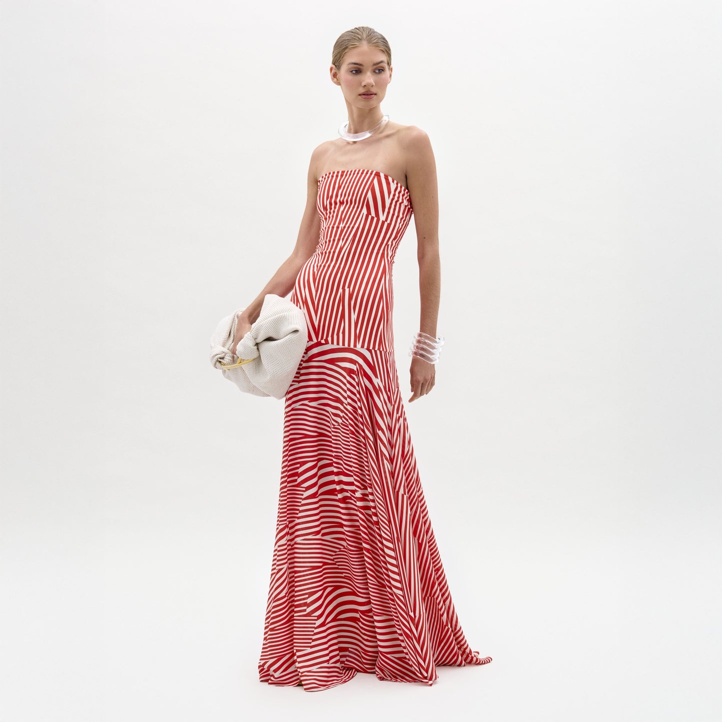 A woman stands against a plain white background in the Muriel Dress—a red and white striped, strapless maxi with a fitted bodice crafted from luxurious Italian fabric. She accessorizes with pearls, a white clutch, and slicked-back hair.