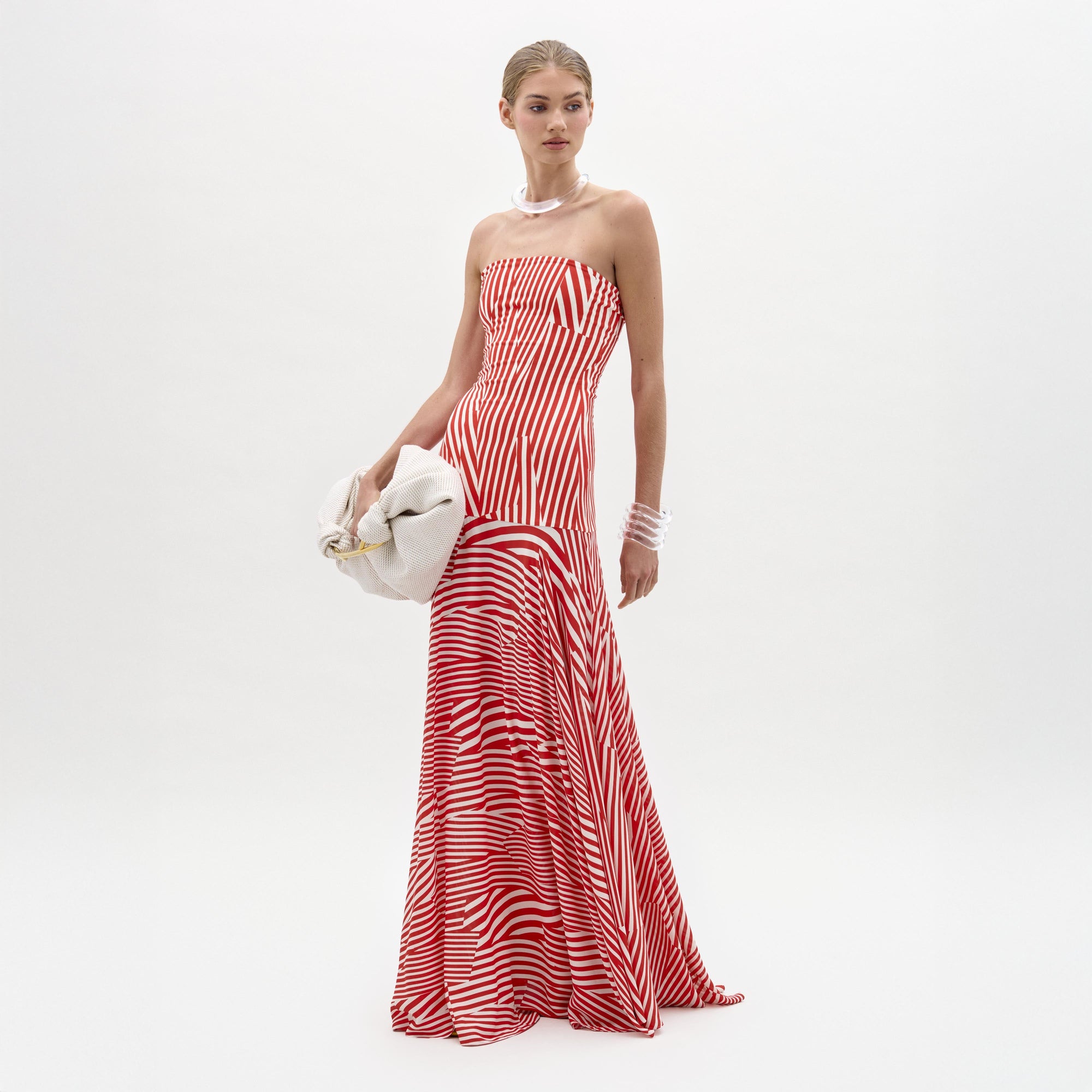 A woman stands against a plain white background in the Muriel Dress—a red and white striped, strapless maxi with a fitted bodice crafted from luxurious Italian fabric. She accessorizes with pearls, a white clutch, and slicked-back hair.