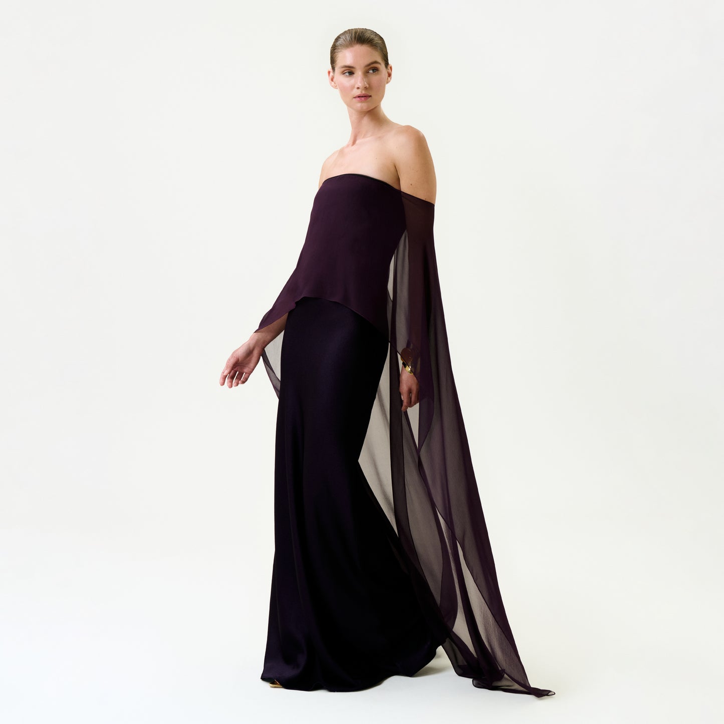 A woman stands against a plain background wearing the Maiya Dress—a strapless dark purple maxi with floor-length sheer sleeves, crafted from sumptuous Italian fabric. She exudes poised, confident elegance.