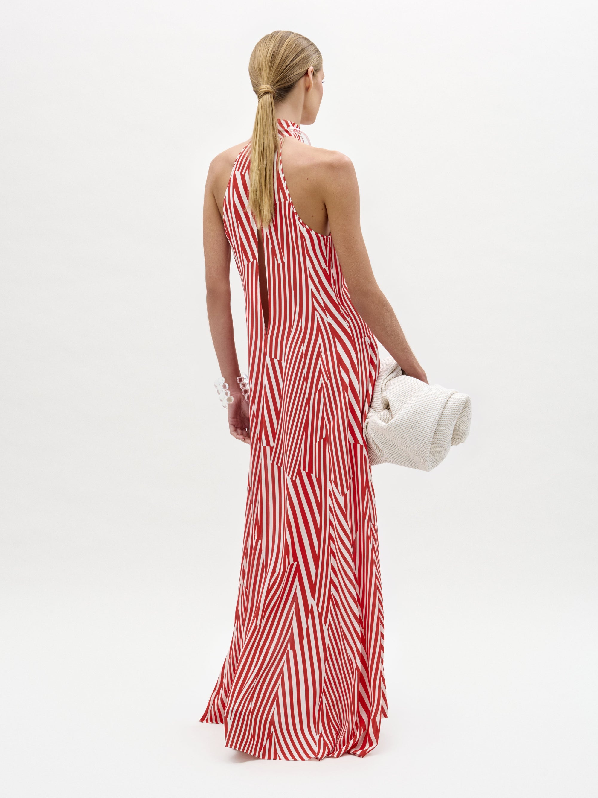 A woman wears the Koa Dress, a red and white striped halter neck maxi with an A-line silhouette, standing against a plain white background while holding a white towel and wearing sandals.