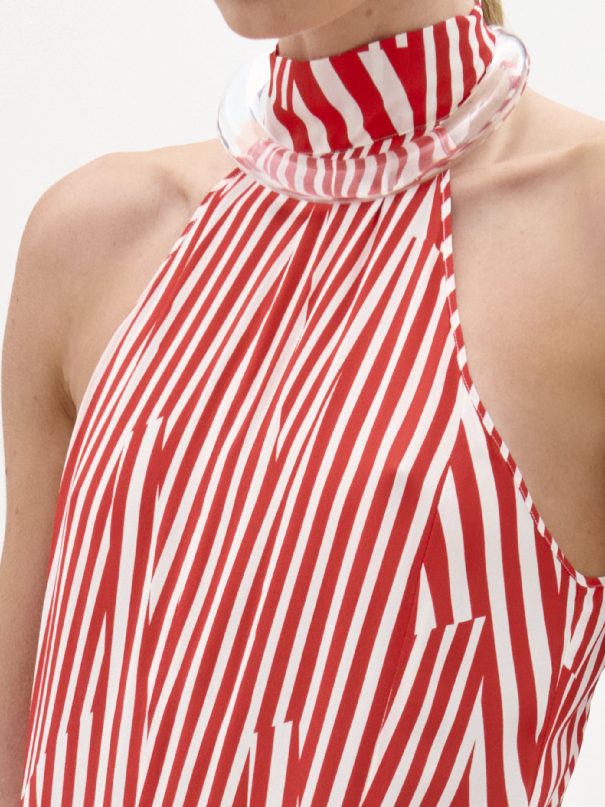 A woman wears the Koa Dress, a red and white striped halter neck maxi with an A-line silhouette, standing against a plain white background while holding a white towel and wearing sandals.